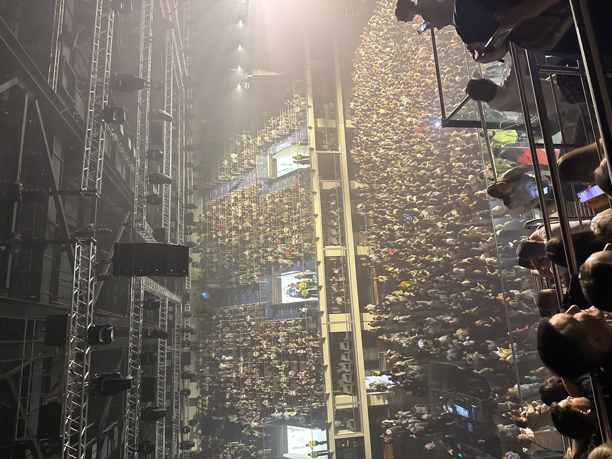 An elevated view of a concert crowd, seemingly taken from a balcony or viewing platform. The crowd fills the stadium floor and tiered seating areas. In the lower right corner, people are looking over a railing, some holding phones. To the left are metal support structures holding lights and speakers. The location is presumably in Ufa, Russia, but no landmarks or other evidence is visible to confirm.