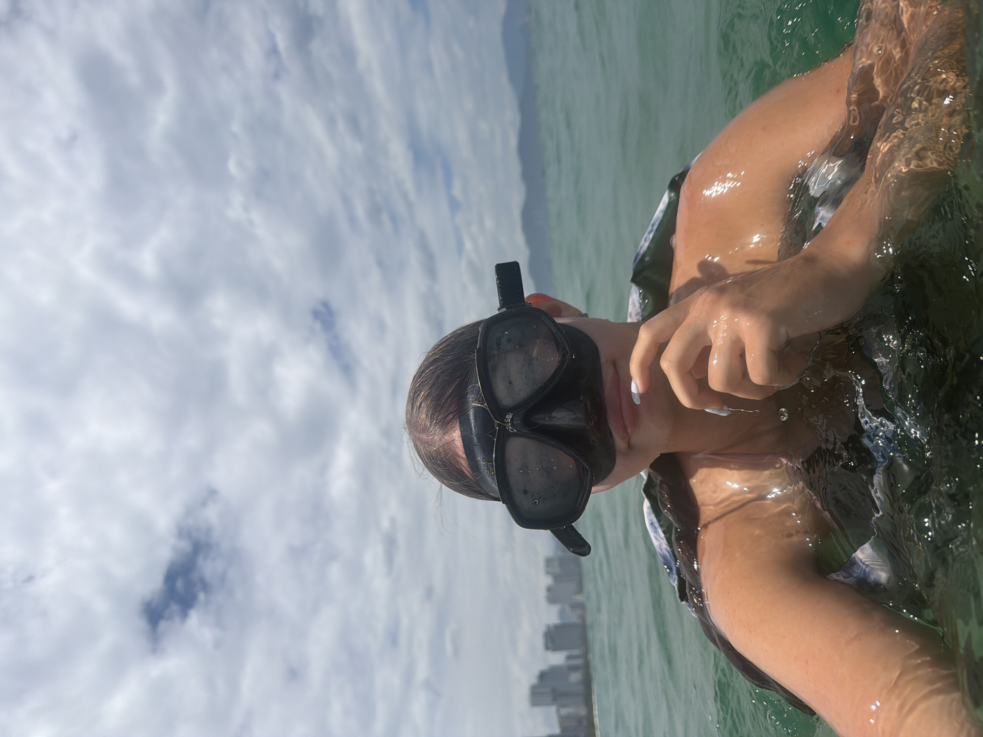 A first-person perspective selfie shows a young person wearing a black diving mask and snorkel in the ocean off the coast of Nha Trang, Vietnam. Buildings of a coastal city are visible in the background under a cloudy sky. The water is green and choppy, with sunlight reflecting off the surface. The person is partially submerged, with their shoulders and arms visible above the water. They are wearing a life vest and a necklace.