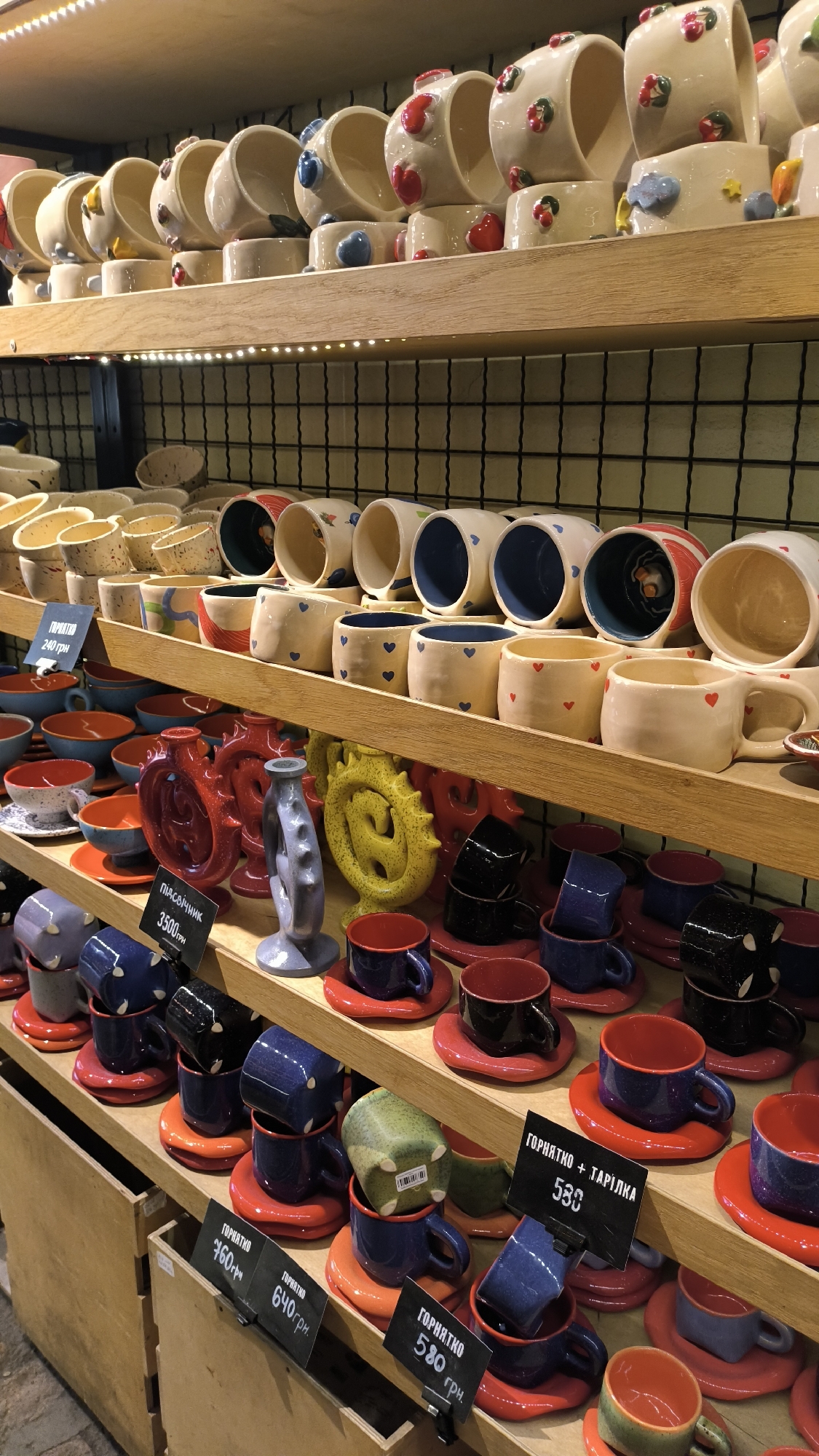 This is a medium shot, eye-level, indoor photograph of several shelves filled with ceramic mugs and other pottery items. The shelves are made of light-colored wood and are stacked vertically. The background consists of a black wire mesh grid.
On the top shelf, there are numerous ceramic mugs with a light cream-colored glaze. Many of these mugs are decorated with colorful heart shapes in red, blue, and green. Some mugs are also adorned with painted blue clouds and yellow stars. The mugs are arranged in rows, with some placed inside others.
The second shelf from the top also features ceramic mugs, many of which are either plain or have blue or red heart patterns. There are also some cylindrical cups visible.
The third shelf displays a variety of ceramic items, including decorative red and blue vessels that resemble vases or jugs. There is also a bright yellow, dragon-like sculpture and a gray, abstract ceramic piece. In front of these, there are rows of red and blue cups and saucers.
The bottom shelf is crowded with various ceramic cups and saucers. There are blue, black, and dark red cups, many of which are stacked. Some of the cups have small tags with prices written in Cyrillic script. For example, one tag reads "ГОРЯЧКО 580 грн.". The overall impression is that of a display of handmade ceramic tableware.