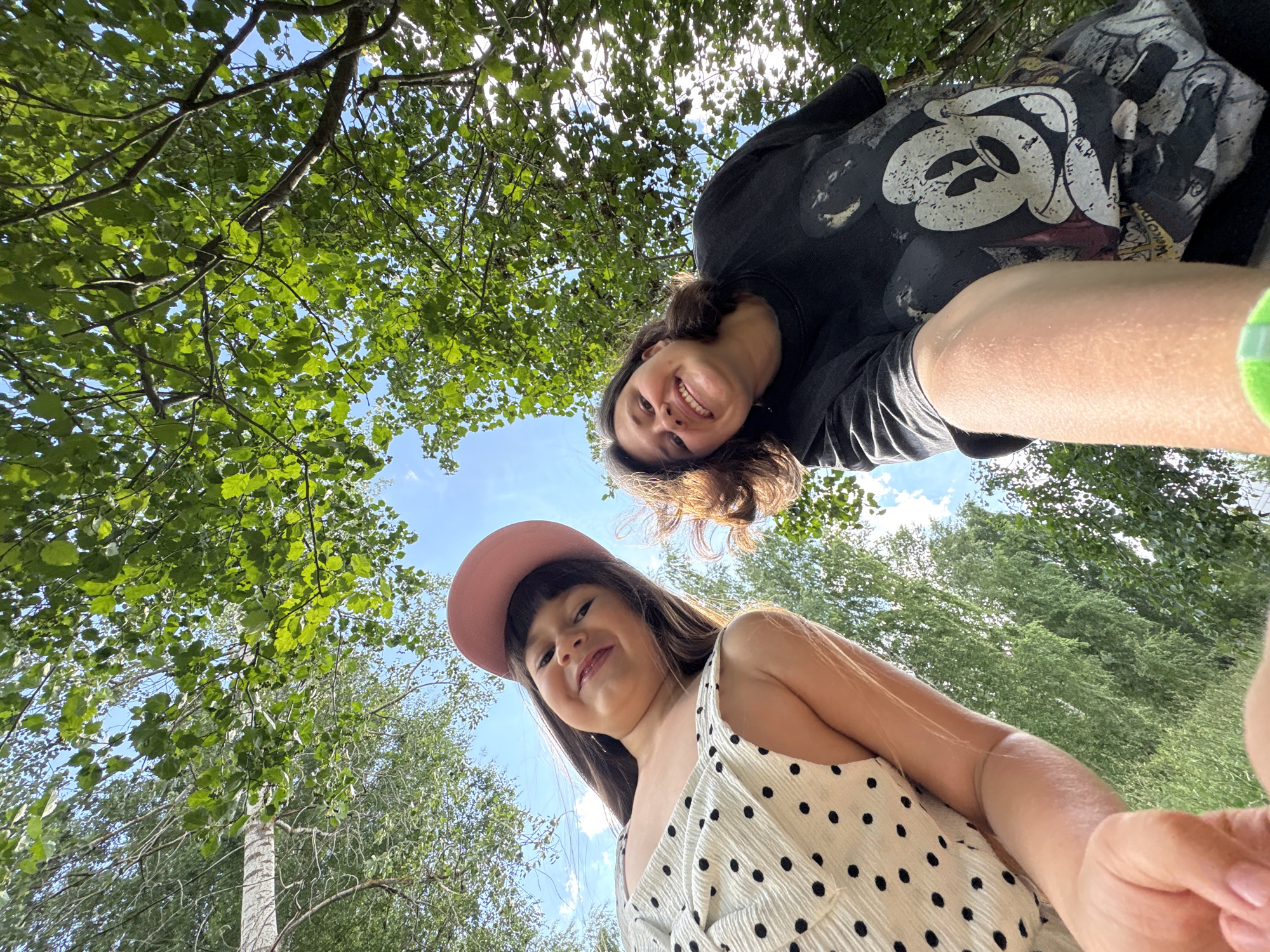 Two people are posing for a selfie outdoors, looking up at the camera from a low angle. The scene is set amongst lush green trees with the sky visible through the branches. The sun is shining brightly, suggesting a clear and sunny day. In the foreground, a young girl wears a pink baseball cap and a white dress with black polka dots. She is smiling at the camera. Behind her and slightly to the left, a woman with long brown hair is also smiling, her face tilted towards the camera. She is wearing a black sweatshirt with a graphic print, possibly of Mickey Mouse. The overall mood appears joyful and relaxed, likely a casual outing in nature. The specific location is identified as Chaikovsky, Russia. There is no visible text in the image.