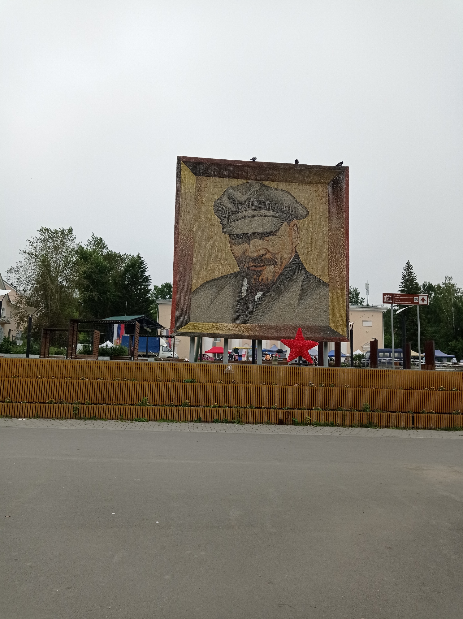 This is a medium-shot, eye-level, outdoor, daytime, overcast photograph of a large mosaic of Vladimir Lenin in the city of Degtyarsk, Russia. The mosaic is framed and depicts Lenin from the chest up, wearing a cap and a jacket. A red star is positioned in front of the mosaic. The mosaic is behind a wooden fence with vertical slats. In the background, there are trees, buildings, tents, a white truck, and street signs. There are also several birds perched on top of the mosaic frame and flying in the sky. The foreground shows an asphalt road.