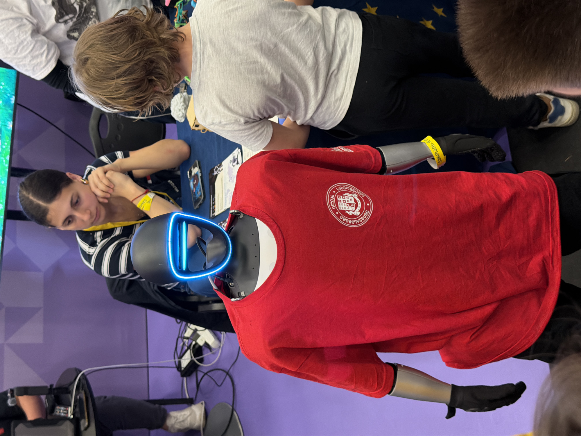 A photograph shows a robot wearing a red t-shirt bearing the Georgian National University logo, and featuring a headset with blue lights. The robot is positioned at a table alongside several individuals, including a woman wearing a yellow wristband. The robot is wearing black gloves. The location is Tbilisi, Georgia.