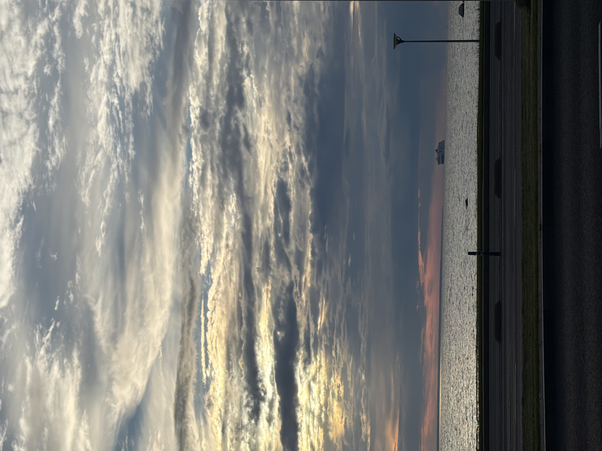 The image depicts a vast, cloudy sky over the Baltic Sea near Tallinn, Estonia. The sky is filled with layers of dramatic, textured clouds, with streaks of sunlight breaking through in the upper left portion of the frame, suggesting either sunrise or sunset. The sea below is a muted grey-blue, with gentle ripples. In the distance, a large ferry is visible on the horizon. The foreground features a paved road with a grassy median and a line of dark bollards. A lamppost stands on the right side of the frame, casting a faint glow. The overall scene is serene and atmospheric, capturing a moment of twilight or dawn over the Estonian coast.