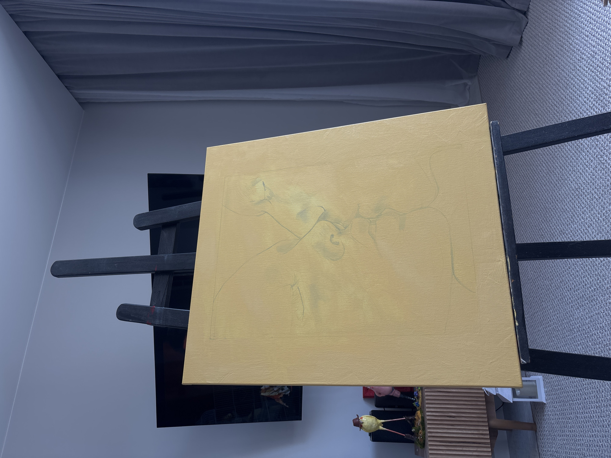 An upright, black easel holds a blank canvas, oriented in portrait mode, which is predominantly primed in a warm, light yellow color. Faint, abstract pencil lines, possibly preliminary sketches, are visible on the canvas surface.
The setting is an indoor room. To the left of the easel, a plain light grey wall is visible. Behind the easel, a dark, reflective surface, likely a television screen, is partially seen. Above, dark grey curtains or drapes are visible, characterized by soft folds, possibly covering a window.
To the right of the easel, a light grey carpet with a subtle textured pattern covers the floor. In the lower right corner, a low-profile, light brown wooden cabinet or furniture piece with horizontal slats is visible. On top of this furniture, several decorative items are placed, including a whimsical figure resembling a bird wearing a hat, and other small, colorful ornaments. A white, rectangular lantern rests on the carpet near the bottom edge of the frame. Part of a person's leg and foot are also visible on the carpet next to the lantern, suggesting someone is present in the room.
The lighting is soft and ambient, casting minimal shadows. No visible text is present. The scene suggests an art studio or a creative space within a home in Krasnoyarsk, Russia, with the canvas poised for an artistic endeavor.