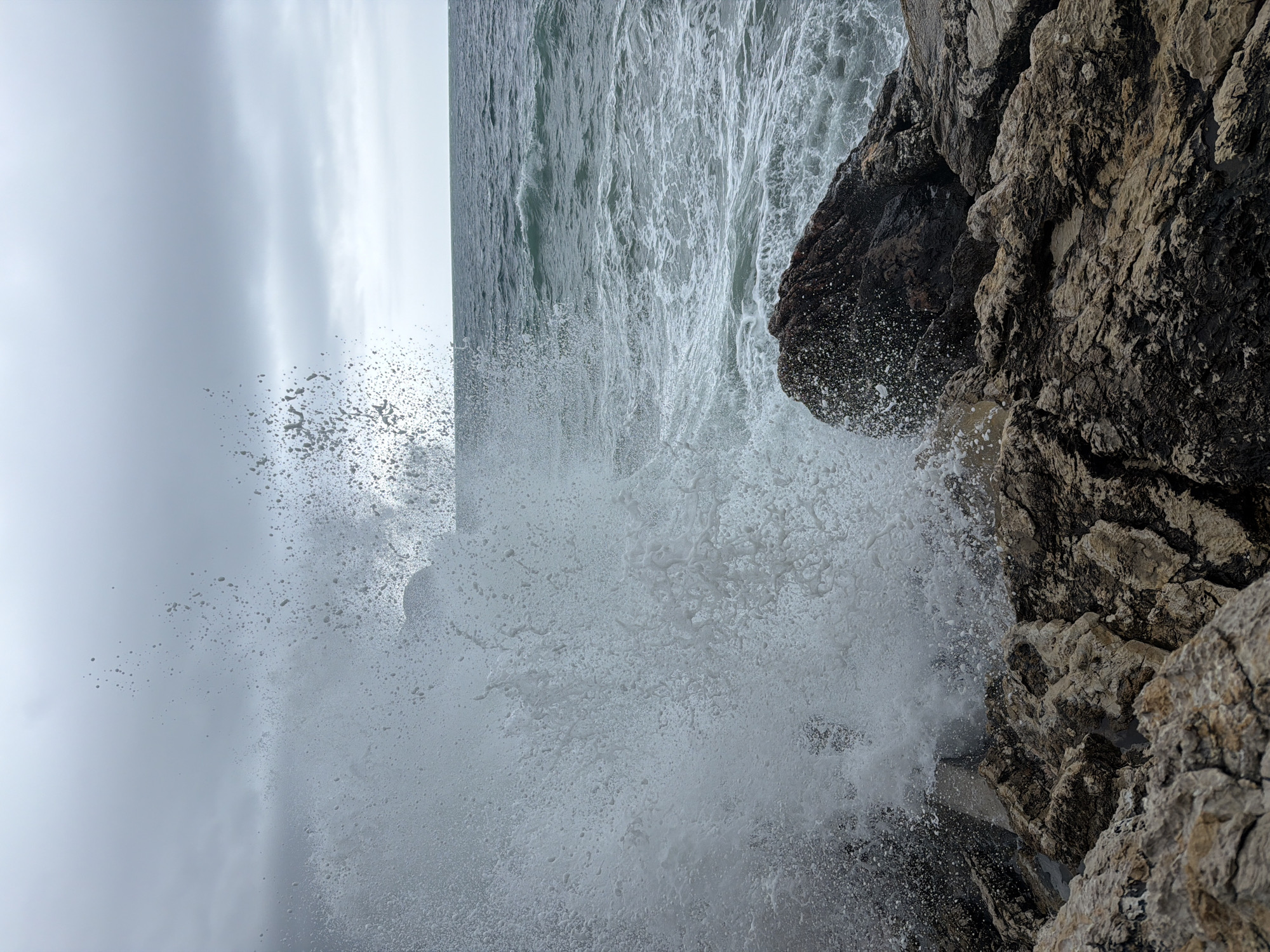 A close-up, slightly tilted, extreme wide-angle shot captures a dynamic scene of the sea crashing against rocky cliffs in Petrovac centar, Montenegro. The rough sea is a dominant presence in the left portion of the frame. The sea spray forms a cloud of droplets near the bottom and dissipates toward the cloudy sky, creating a blurred visual boundary. The rocky cliffs occupy the right portion of the image, their texture and crevices sharply defined. The color palette is muted, dominated by gray, white, and brownish hues reflecting the overcast weather. The scene portrays the raw power of nature and the interaction of water and rock.