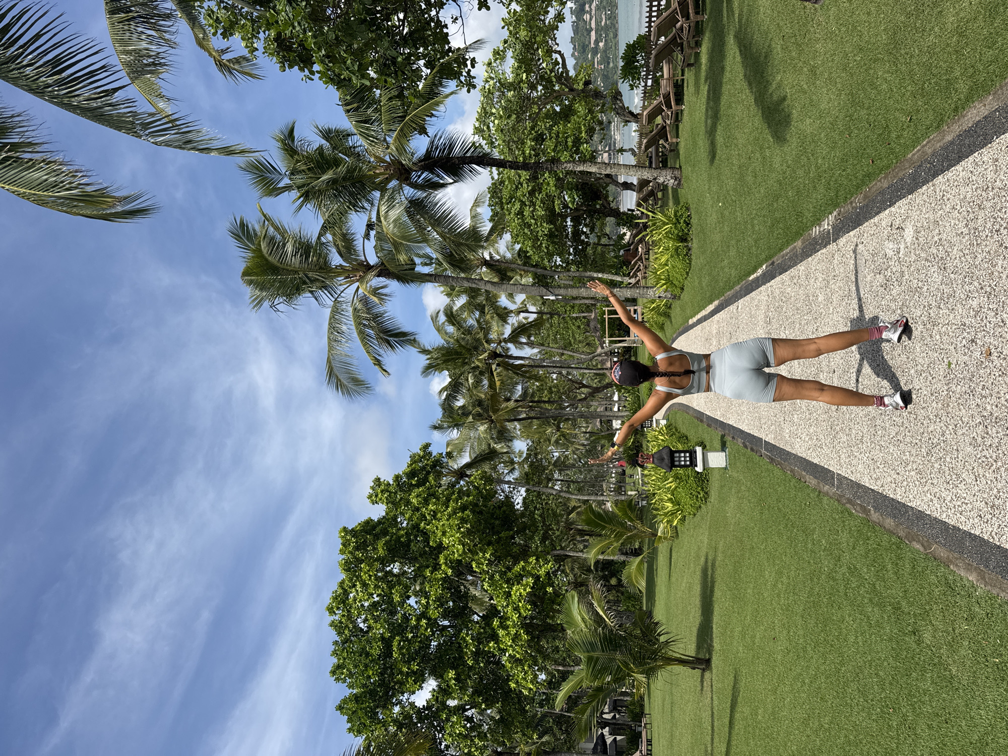 A person stands with arms outstretched on a pathway in Kuta Selatan, Indonesia. The path, composed of small stones, runs through a grassy area lined with palm trees and other greenery. The person is wearing athletic clothing and sneakers. In the background, a body of water and buildings are visible. The sky is blue with scattered clouds.