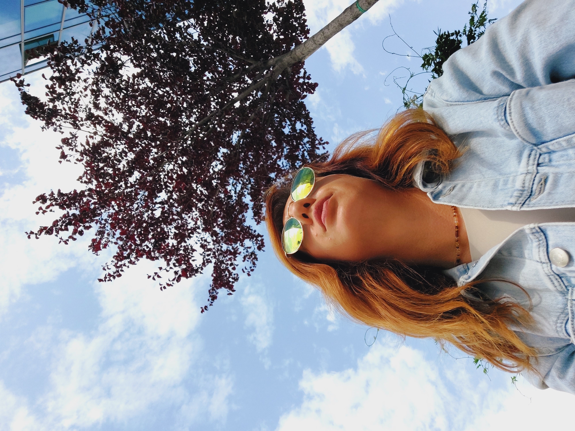 A low-angle, medium shot captures a woman with reddish-blonde hair wearing mirrored sunglasses and a light blue denim jacket. She is looking upwards. Behind her, a tree with dark red leaves is visible against a bright blue sky with scattered white clouds. To the upper right, a corner of a modern building with reflective blue glass windows can be seen. The scene appears to be an outdoor urban environment.