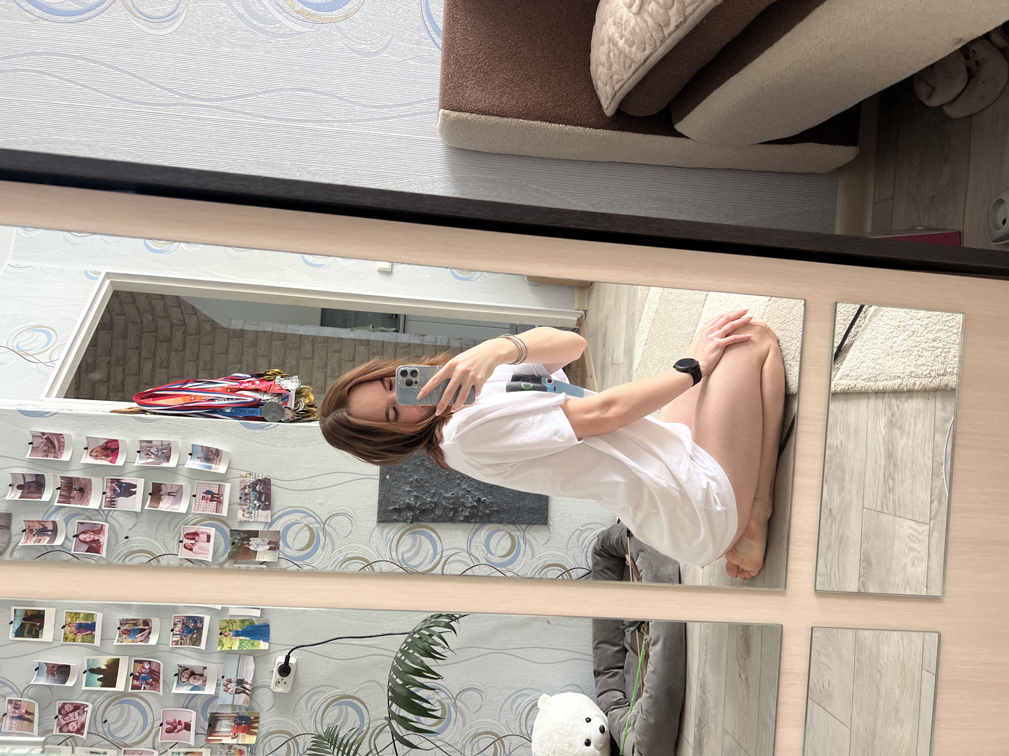 A young woman is taking a selfie in a mirror made of four square panels, presumably in a residence in Chaikovsky, Russia. She is wearing a white t-shirt and is seated on the floor. In the background, there is a wall decorated with photos and medals, along with a couch and plant. A black-framed mirror surrounds the composition.