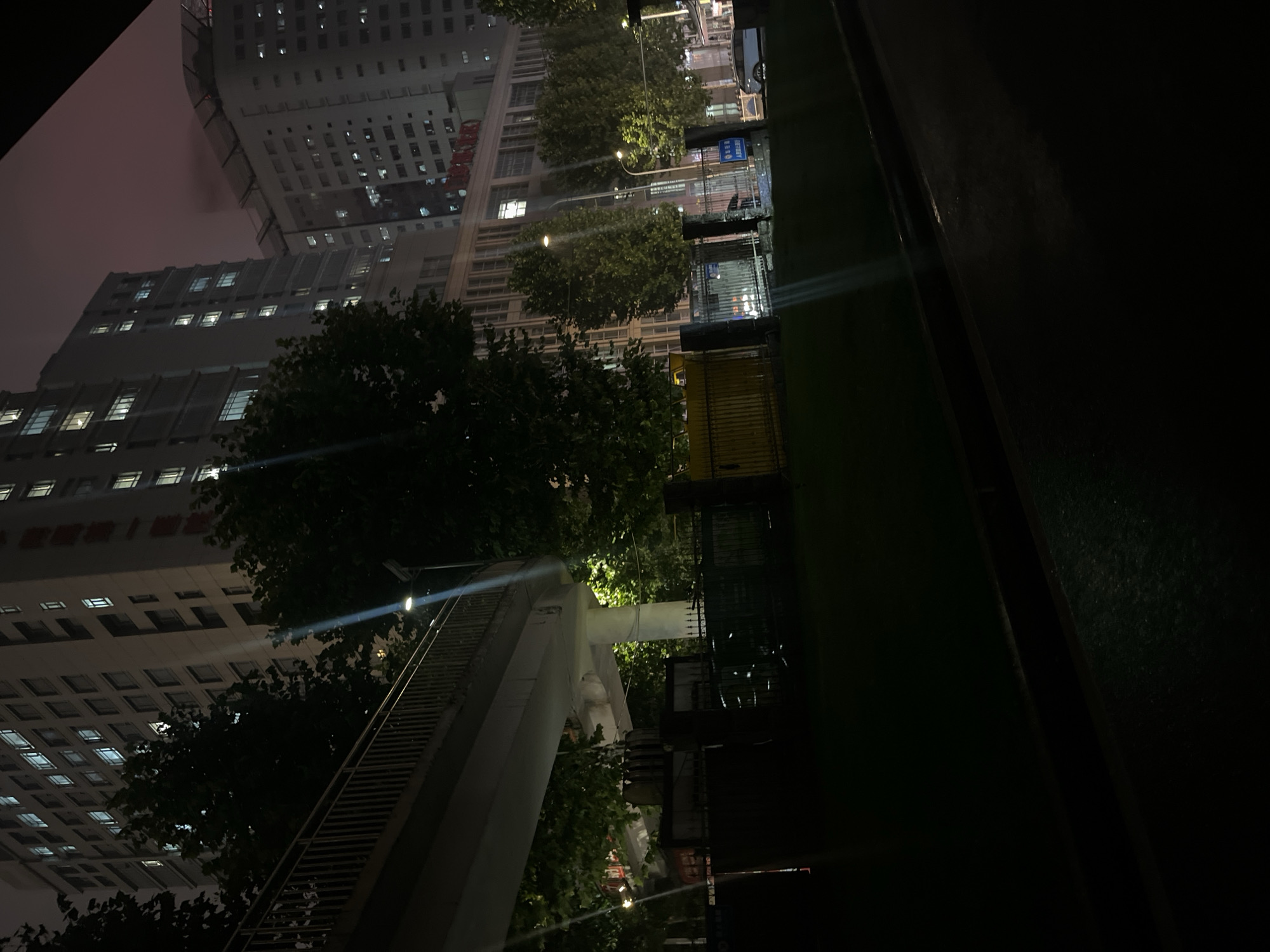 This image depicts an urban nighttime scene in Yuzhong District, Chongqing, China, specifically in the vicinity of Chongqing First Hospital, identified by the legible red Chinese characters "重庆第一医院" on a building facade.
The scene is dominated by multiple high-rise buildings under a dark, reddish-purple sky, illuminated by numerous interior lights from windows. The primary building on the left is a tall, multi-story structure with rectangular windows. To its right, another tall building with a distinctive rounded top, possibly housing a helipad, is visible, also with illuminated windows. Beneath and in front of these, a lower, multi-story building features the aforementioned "Chongqing First Hospital" signage.
In the midground, a concrete pedestrian overpass with metal railings curves across the frame from left to right, supported by a single visible concrete pillar. Below the overpass, a dense arrangement of dark trees and foliage is present, with some sections brightly lit by streetlights.
The foreground consists of a dark, paved surface, possibly a road or walkway, exhibiting reflections consistent with a wet surface. A dark metal fence runs horizontally across the midground, separating the foreground from the area beneath the overpass and the buildings. Behind this fence, a section of yellow fencing is discernible.
Several streetlights are active, casting strong, visible beams of light, particularly near the overpass and illuminating portions of the trees and ground. On the right side of the paved area, a white car is parked, and a single individual holding an umbrella is faintly visible near it, suggesting recent or ongoing precipitation. No other individuals or active interactions are clearly identifiable.