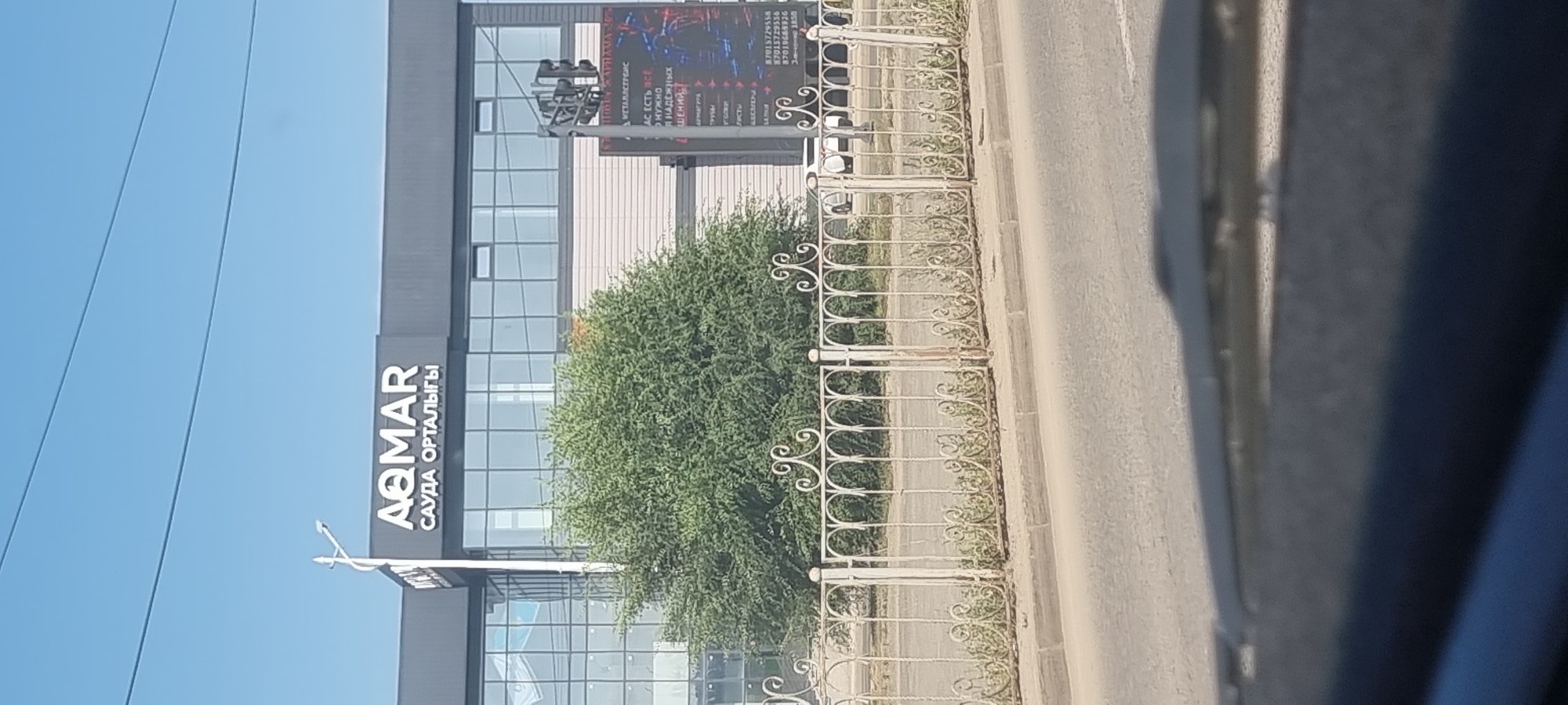 The image shows a street view in Aktobe, Kazakhstan. A modern building with large windows and the prominent sign "AQMAR SAUDA ORTALIGЫ" is visible. In front of the building, a large green tree stands. A white ornamental fence separates the building from the street. To the right of the building, a digital billboard displays text and images, which are partially legible. The street in the foreground appears to be a road with some vegetation growing along the curb. The overall scene suggests a commercial or business area.
