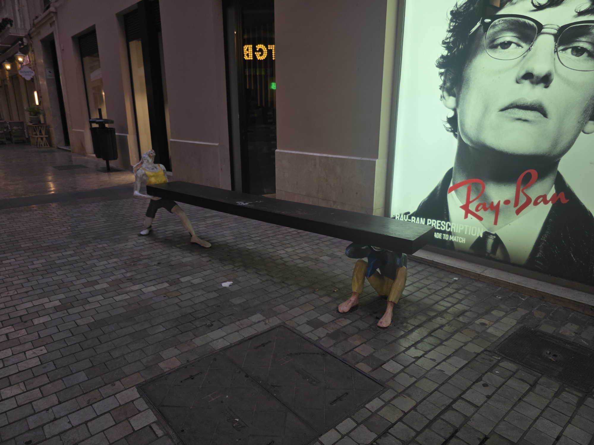 In Málaga, Spain, an outdoor scene unfolds on a cobblestone street at dusk. Two life-sized mannequins are positioned to support a long, dark wooden bench, creating an artistic installation. The mannequin on the left, with a pale complexion, wears a yellow top and dark shorts, and is seen from the knees up, holding the bench with its hands. The mannequin on the right, also visible from the knees up, has yellowish-tan skin and wears blue shorts over yellow leggings. It is crouched down, supporting the bench with its arms.
The background features the facade of a building with large windows, some illuminated, hinting at shops or businesses. A prominent advertisement for Ray-Ban glasses, featuring a close-up of a person wearing glasses, is visible on the right side of the frame. The street is paved with grey cobblestones, and a manhole cover is visible in the foreground. The lighting suggests the time is evening, with artificial lights from the building and the advertisement illuminating the scene.