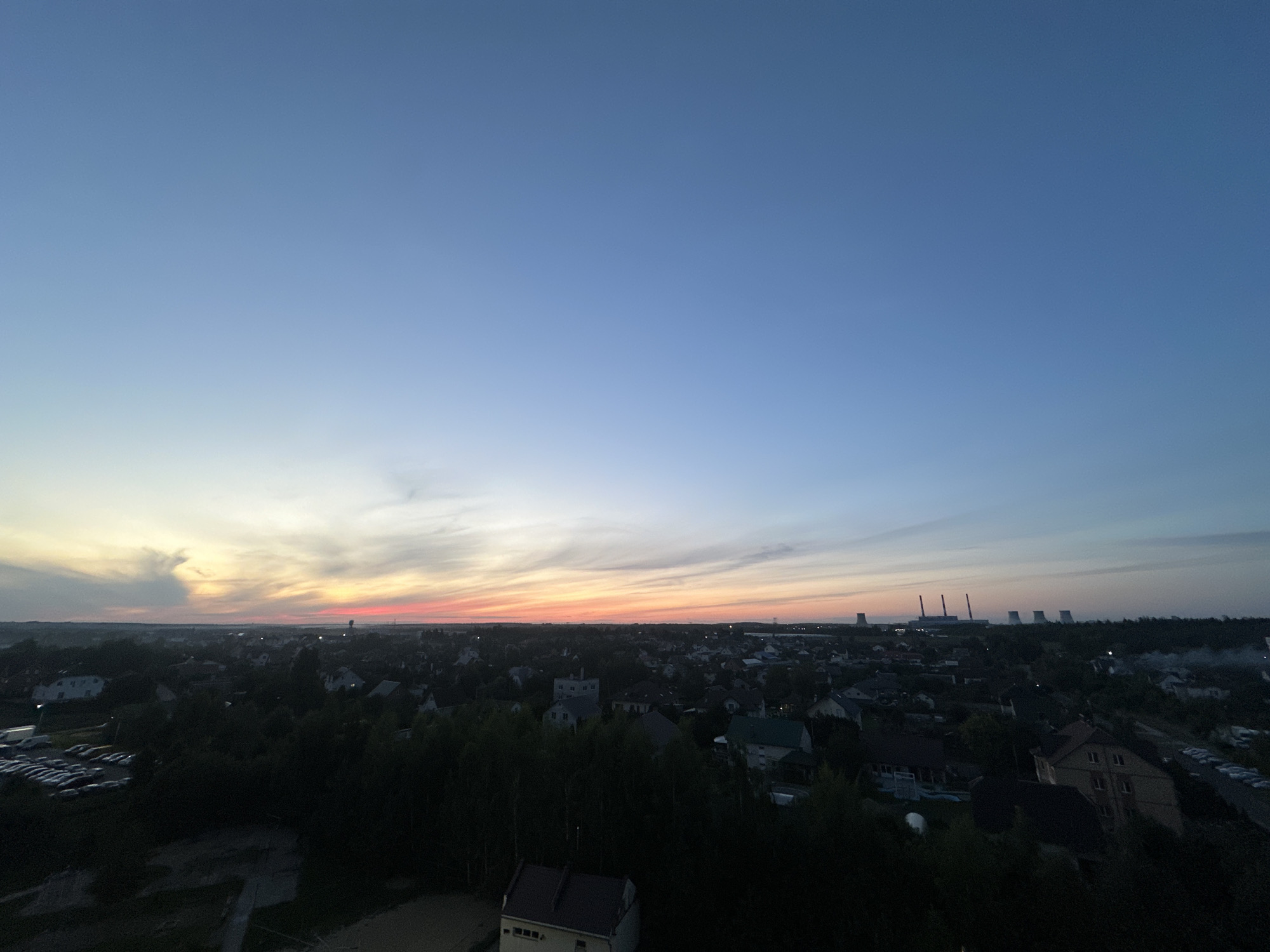 An aerial wide-angle shot captures the twilight sky over the Шчомысліцкі сельскі Савет in Belarus. The sky transitions from a deep blue overhead to hues of pink, orange, and yellow near the horizon, where a silhouetted treeline marks the edge of a suburban landscape. Numerous houses with dark roofs are visible, interspersed with trees. To the left, a parking lot filled with white cars is partially obscured by dark foliage in the foreground. In the distance, several tall, industrial structures, possibly power plant cooling towers, punctuate the skyline. The overall scene suggests a quiet evening in a residential area with industrial elements on the periphery.