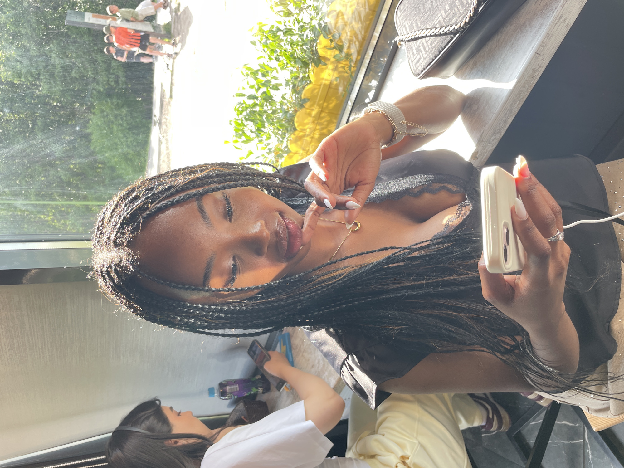 A Black woman with braided hair, wearing a black top, looks down at her white smartphone. Her right hand is positioned near her chin, and her left hand holds the phone. She is wearing a necklace and a bracelet. In the background, another woman in a white shirt is visible, holding a phone. Outside the window, a group of children are playing. The scene appears to be set indoors, near a window that lets in sunlight.