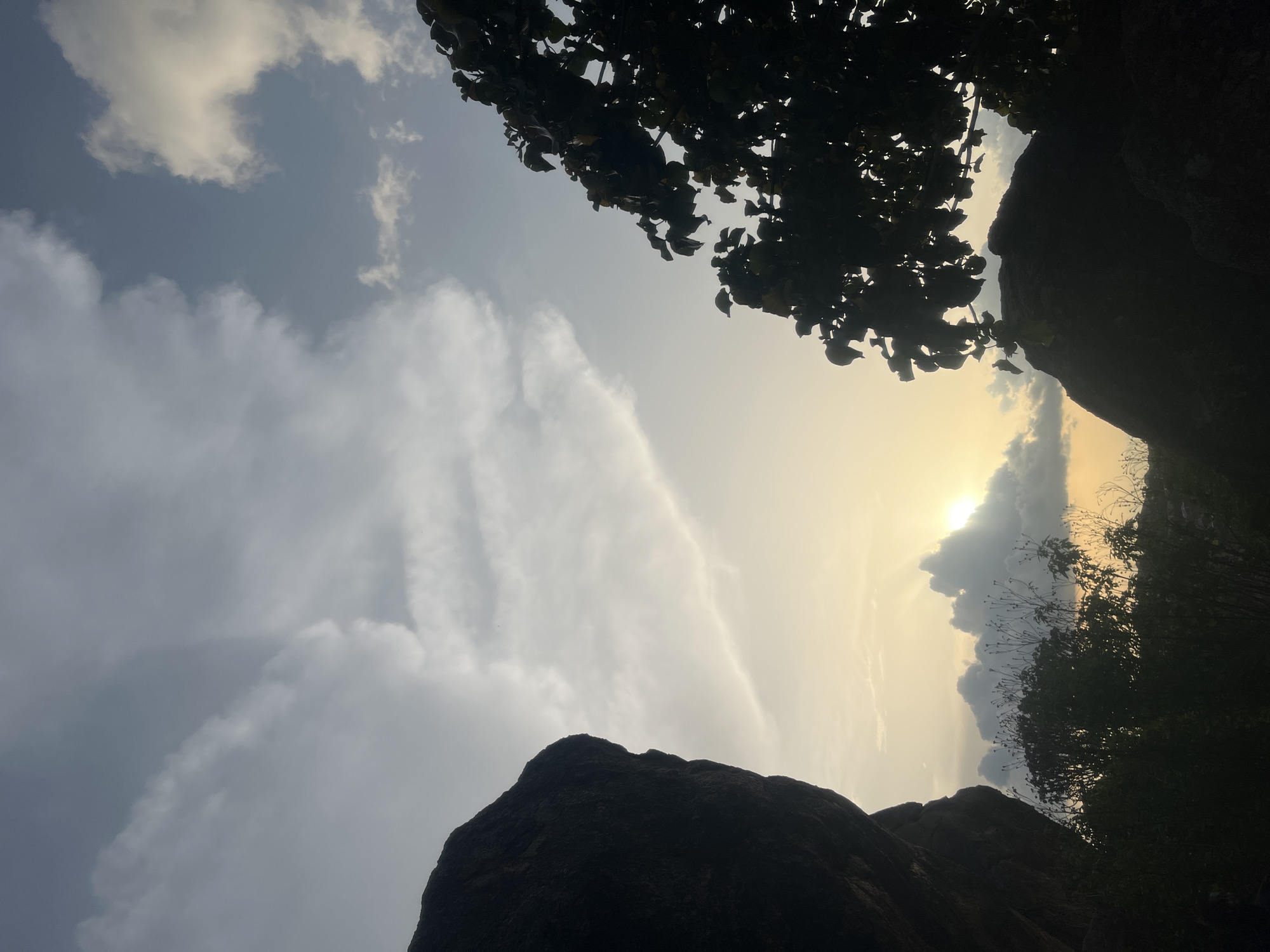 The image displays a natural outdoor scene at either sunrise or sunset, within the city of Jos, Nigeria. The upper two-thirds of the frame are occupied by a sky exhibiting a gradient from a warm, yellow-orange hue near the horizon in the lower-central area to cooler tones of grey and pale blue in the upper regions. The sun is visible in the lower-center-right, partially obscured by dense, dark clouds, from which distinct crepuscular rays project upwards. A variety of cloud formations, ranging from elongated, wispy structures to more voluminous, dark masses, are distributed across the sky.
The left side of the frame is dominated by a large, dark rock formation, presented in complete silhouette against the brighter sky. Its rugged edge is prominent. The right side of the image features a silhouetted tree, extending from the top to the mid-right, with discernible individual leaves. The lower third of the scene, encompassing the foreground and midground, consists of a dense arrangement of dark, silhouetted vegetation, including bushes and smaller trees, interspersed with additional rocky outcrops. No human figures, animals, or man-made structures are visible within the frame. The scene portrays a rugged, vegetated landscape illuminated by the low angle of the sun.
