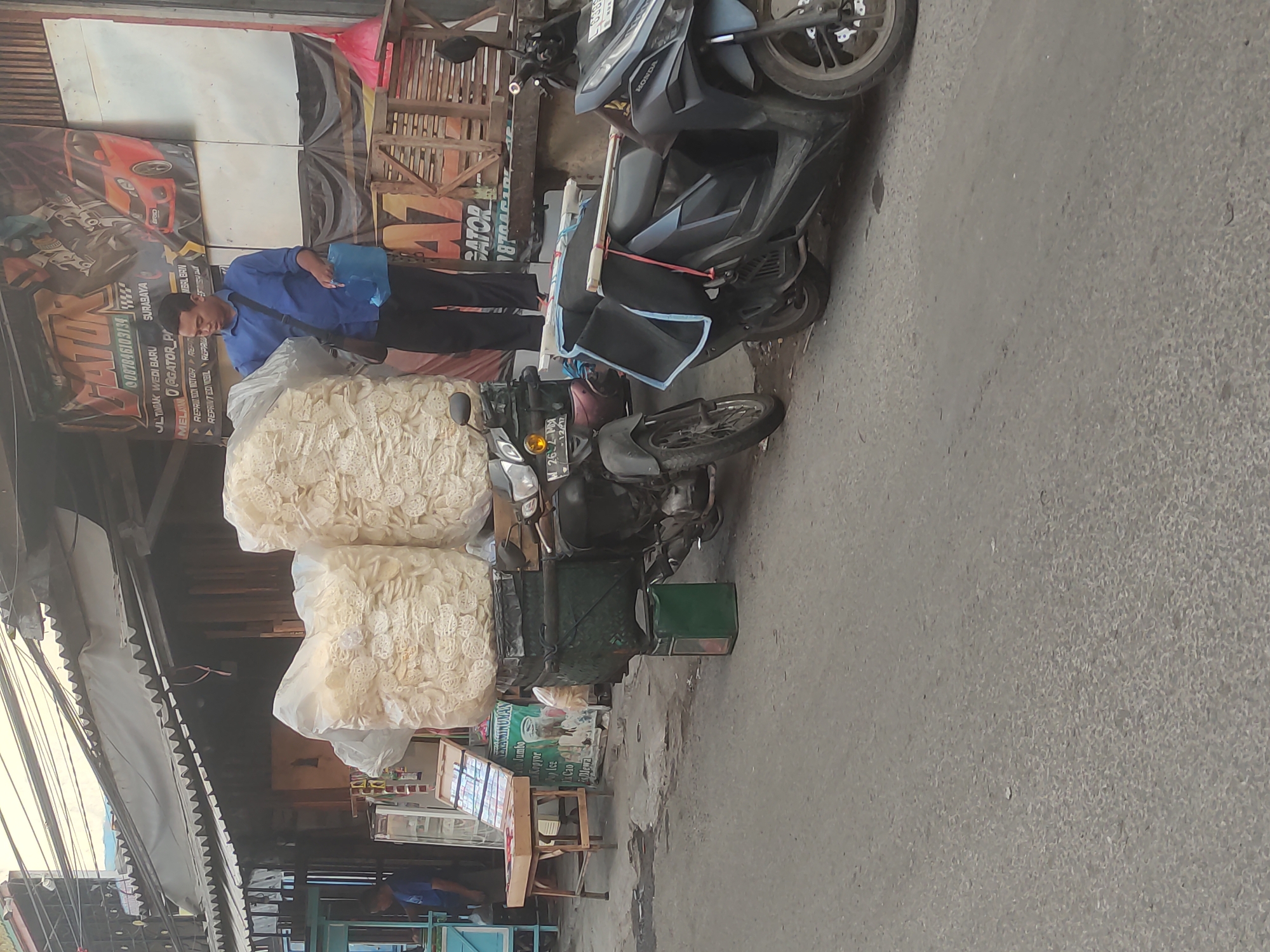 In Surabaya, Indonesia, a man in a blue shirt and black pants stands next to a motorcycle laden with large, clear plastic bags filled with light-colored crackers or snacks. The motorcycle is parked on a paved street in front of a building with a corrugated metal awning. Behind the man, a banner displays "GATOR" with contact information and imagery of a car. Another motorcycle is parked to the left, and a small wooden table with various items on display is to the far left. A green bin is placed beneath the cargo on the first motorcycle. The environment appears to be a bustling urban street.