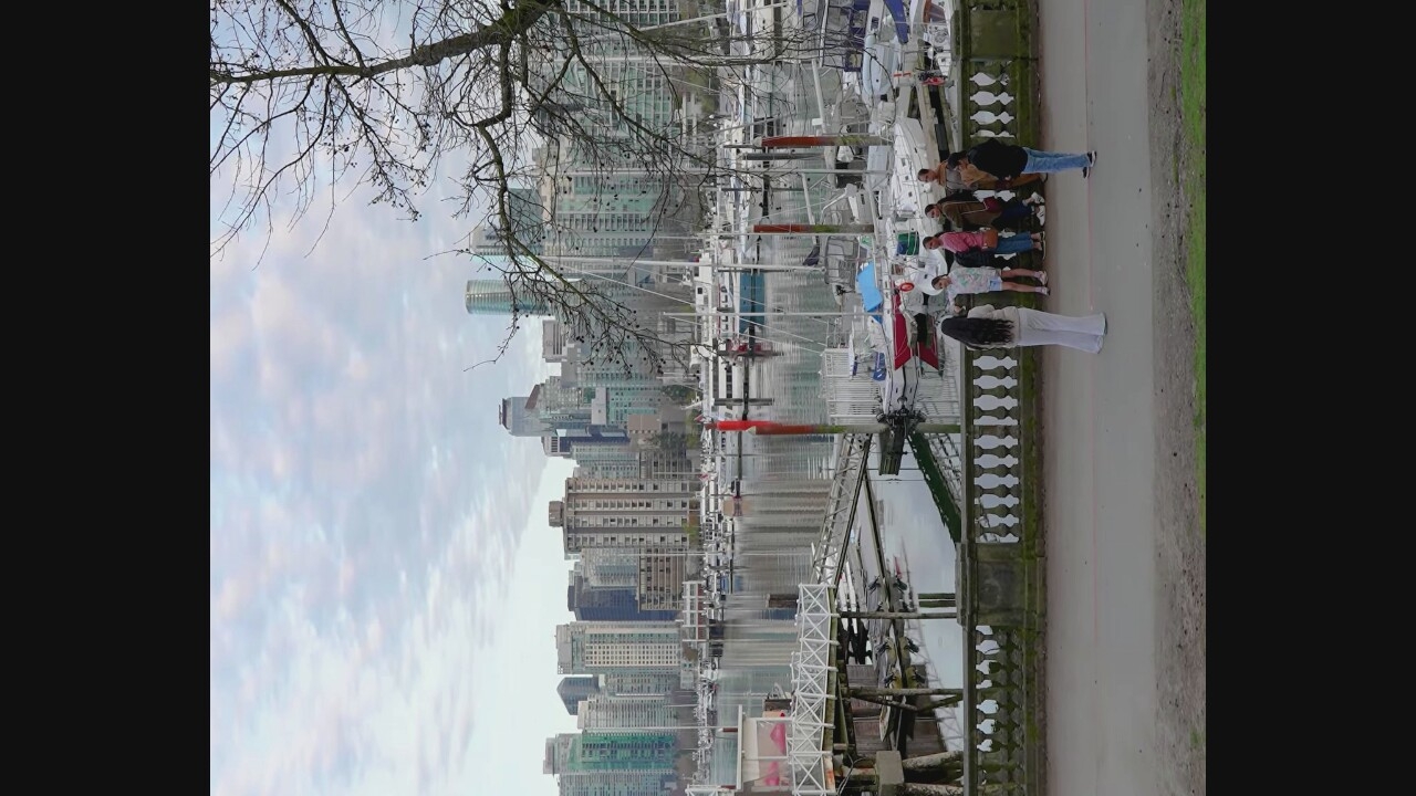 The image depicts a scene at Coal Harbour Marina in Vancouver, Canada. In the foreground, a woman in white pants and a beige top stands with her back to the camera, facing a group of five people. This group includes two women in the center, one wearing a pink shirt and the other a brown jacket, both looking towards the woman in white. To their right, another woman in a darker jacket and jeans is visible, and to the far right, a man in a dark jacket and blue jeans is looking at the group.
Behind the people is a stone balustrade and a pathway. Beyond the pathway, the Coal Harbour Marina is visible, filled with numerous sailboats and motorboats docked in the water. A white wooden pier with ramps extends into the marina. The background is dominated by the Vancouver cityscape, featuring a dense collection of modern high-rise buildings. The sky is overcast with scattered clouds, and a bare tree branch with dark berries is visible in the upper right portion of the frame. The water reflects the buildings and the sky.