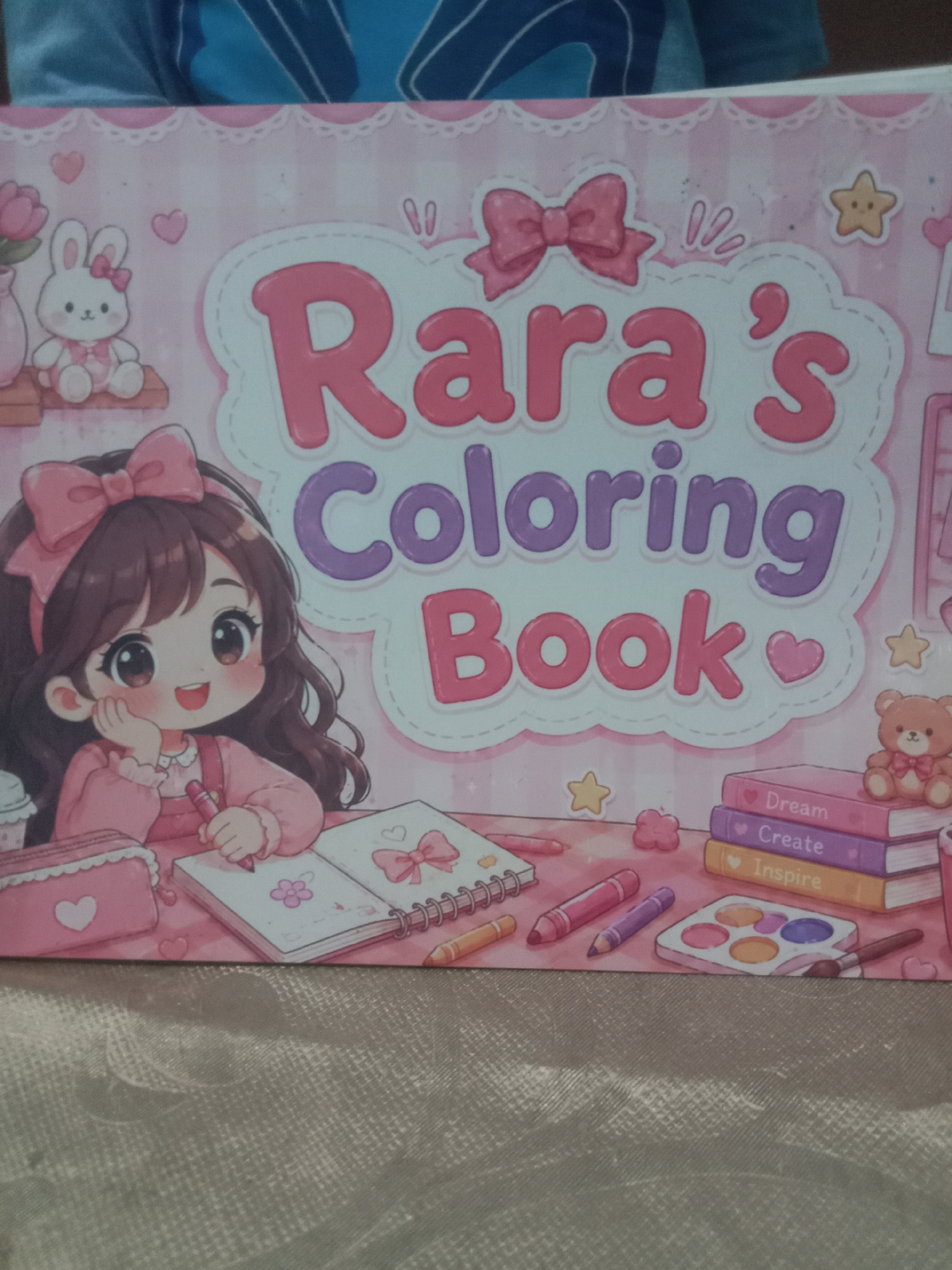 The image is a close-up of the cover of a coloring book titled "Rara's Coloring Book." The cover features a cartoon illustration of a young girl with dark hair and large eyes, wearing a pink dress and a pink bow in her hair. She is depicted drawing in a notebook with a pink crayon. Various art supplies, including crayons, a watercolor palette, and colored pencils, are scattered around her. A stack of books with titles "Dream," "Create," and "Inspire" is visible, along with a small teddy bear. The background is a soft pink with decorative elements like hearts, bows, and stars. The coloring book is placed on a textured surface, possibly a table or floor. The image is taken from a slightly elevated angle, focusing on the coloring book cover. There is no information about the city of Kedungsari, Indonesia, in the image or its contents.