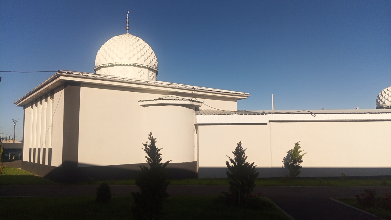 The image shows a mosque with a white dome and minaret under a clear blue sky in Balykchy, Kyrgyzstan. The mosque is a rectangular building with a light-colored facade. In front of the mosque, there are three small evergreen trees. The ground in front of the mosque is covered with grass. The scene appears to be a sunny day.