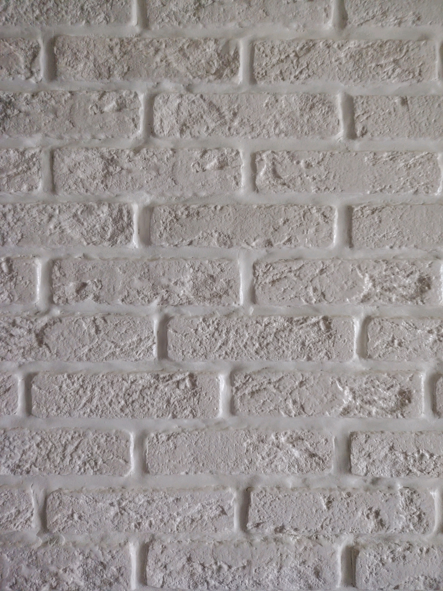 The image displays a close-up, vertical view of a white brick wall. The bricks are rectangular and have a textured, somewhat rough surface. The mortar between the bricks is also white and appears slightly raised, creating a distinct visual separation. The lighting suggests a slightly diffused light source, casting subtle shadows that highlight the texture of the bricks and mortar. There are no people, animals, or other objects visible in the frame, and no specific environmental context is provided beyond the wall itself. The image does not contain any information that would allow for its placement within the city of Chaikovsky, Russia, or any other specific geographical location.