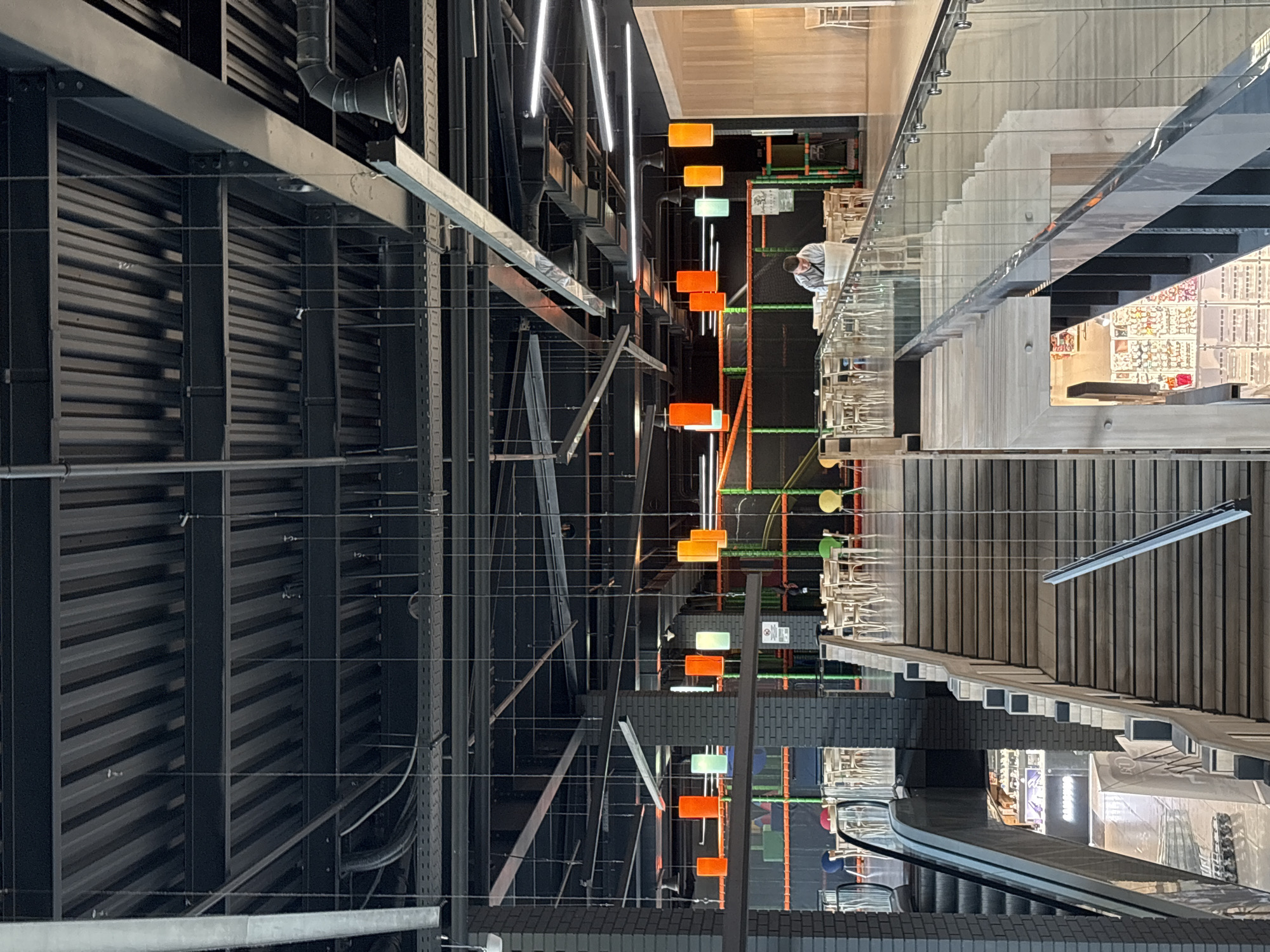 From an elevated vantage point, the image presents an abstract, orthogonal view of the interior of a building in Hatne, Ukraine. The left side of the image is dominated by the building's infrastructure: a dense network of black metal beams, pipes, and wiring against a background of corrugated metal ceiling. The right side displays a multi-level interior, likely a commercial space. This includes a staircase with wooden steps and a glass railing, leading down to a retail area with displayed merchandise. On another level there are cafe style tables and chairs. Between these two distinct sections is a suspended, decorative element consisting of vertical rows of orange and green geometric shapes. The scene appears vacant.