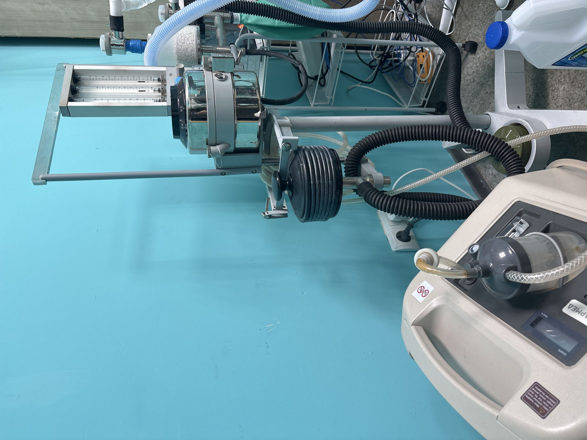 The image depicts medical equipment, specifically an anesthesia machine and a medical suction device. The anesthesia machine features flowmeters, a vaporizer, and corrugated breathing circuits. The suction device is a portable unit with a canister and control panel. The location appears to be a medical setting with light blue walls. There are no people visible in the image. It is impossible to determine if this is Izhevsk, Russia, or anywhere else.