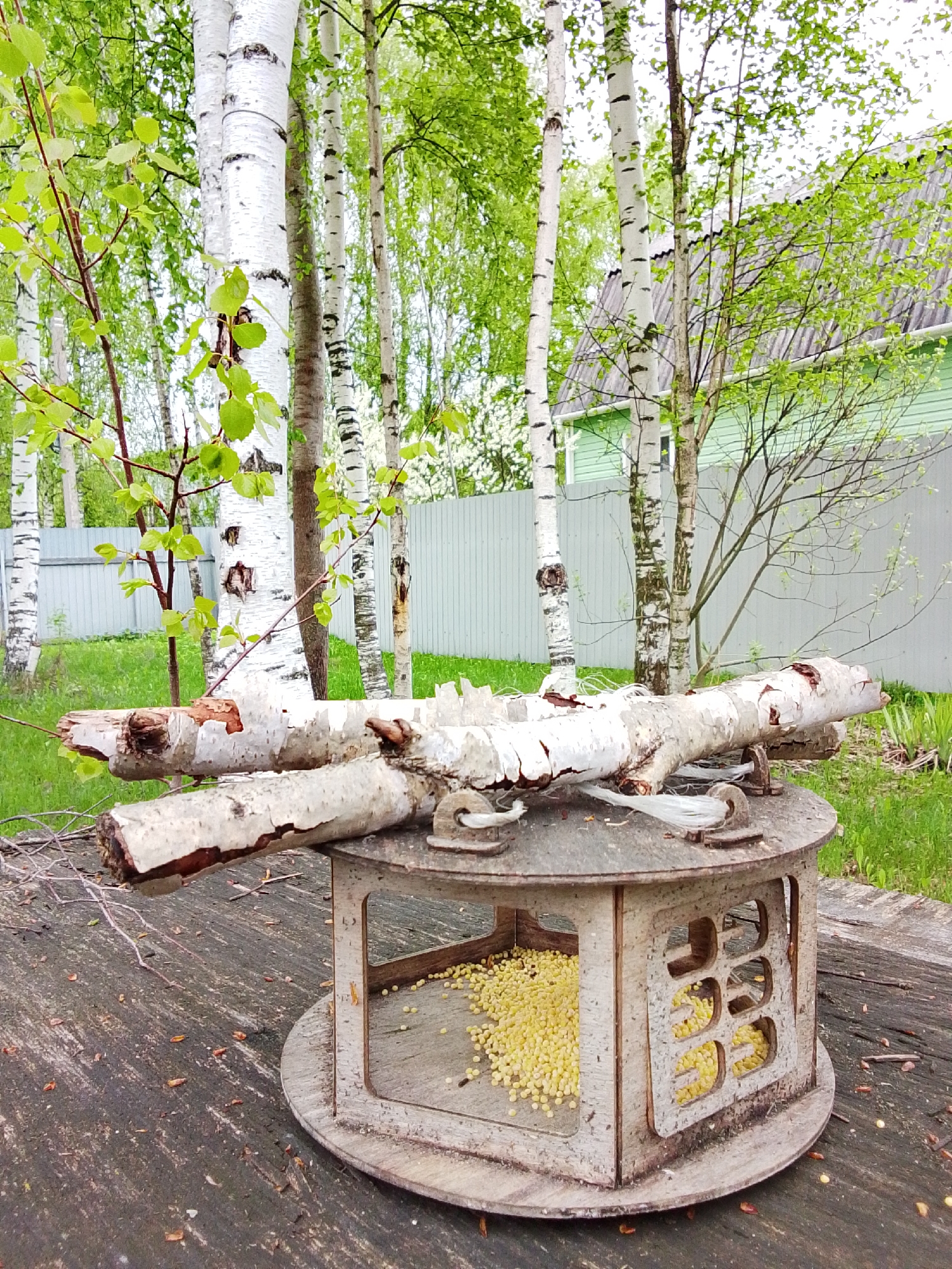 The image shows a rustic bird feeder placed on a tree stump in a grassy area with birch trees in the background. The bird feeder is made of wood and contains yellow birdseed. A birch branch rests on top of the feeder. In the background, there is a fence and a light green building. The scene appears to be in a residential area with natural landscaping. While the image doesn't explicitly state the location, the presence of birch trees and the type of construction are consistent with environments in Russia.