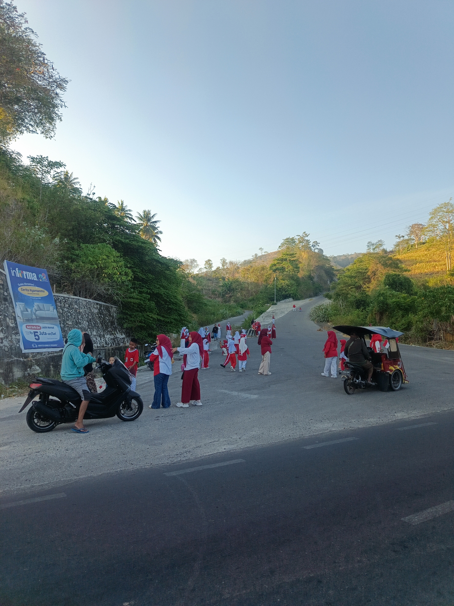A group of people, mostly children wearing red and white uniforms, are gathered on a paved road in a hilly, vegetated area. The scene appears to be in the city of Gorontalo, Indonesia. The road curves upwards into the distance, with lush green trees and vegetation lining the sides. A large billboard for "Informa" is visible on the left, advertising furniture. A person on a black motorcycle, wearing a blue hoodie and shorts, is on the left side of the road. Further up the road, a three-wheeled vehicle is parked. The sky is clear and blue, suggesting it is daytime. The overall impression is of an outdoor gathering or activity taking place on a sunny day in a semi-rural or suburban part of Gorontalo.