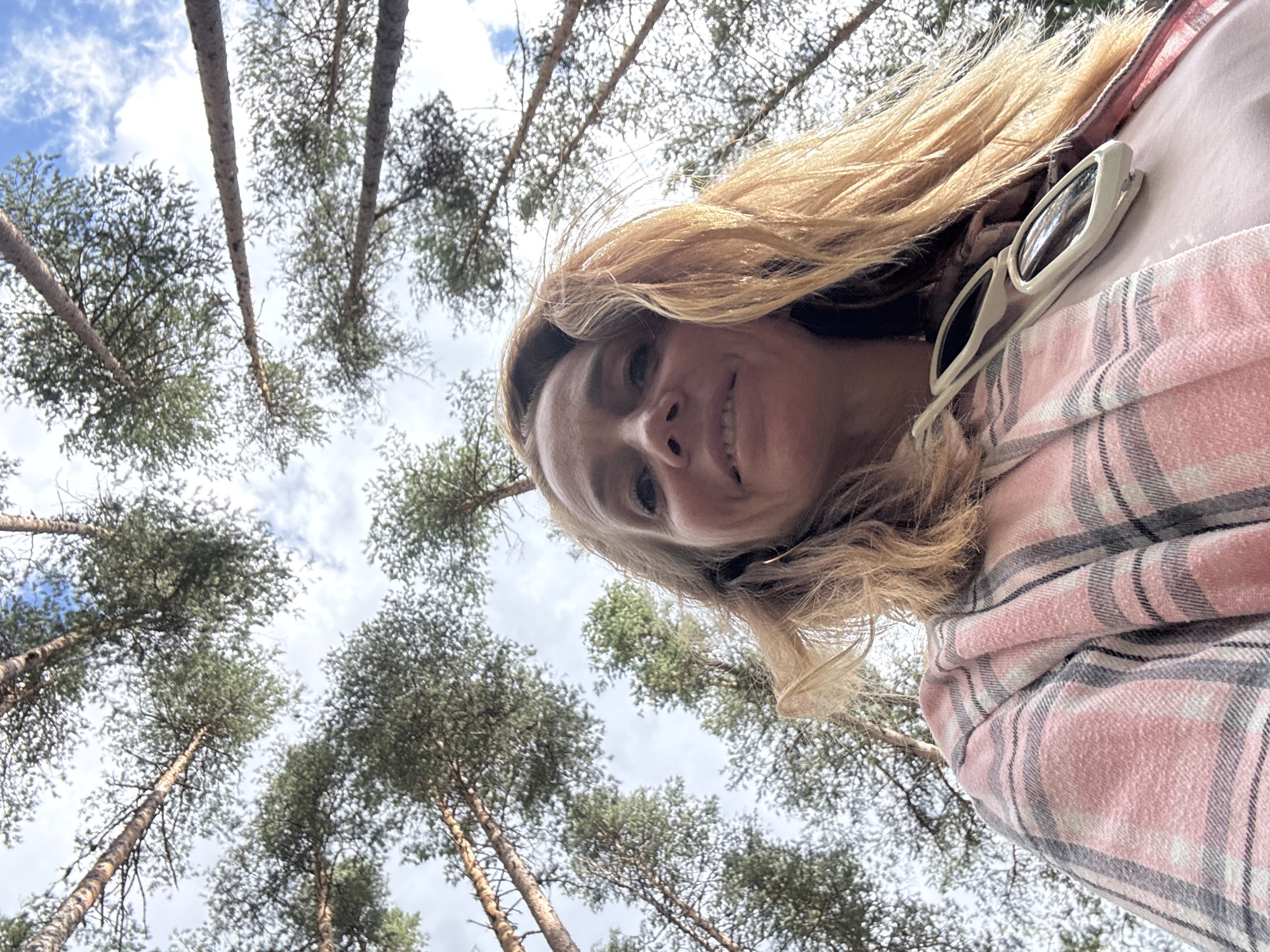This is a low-angle, full-body selfie taken outdoors, looking up at the sky and trees. A woman with blonde hair, wearing a pink and grey plaid shirt and a pink t-shirt, is the primary subject. She has sunglasses hooked onto her shirt. The background consists of tall pine trees reaching towards a cloudy blue sky, suggesting a forest or wooded area. The image does not provide any information that would allow for its localization in Ikhala, Russia, or any other specific city.