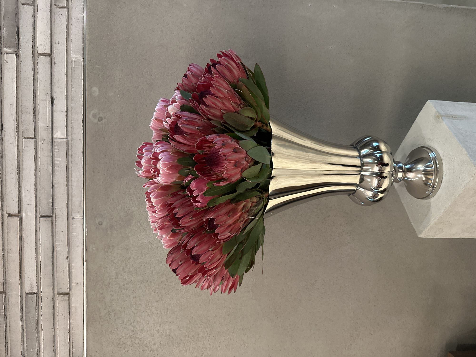 A vase of pink protea flowers sits atop a white column next to a textured wall. The image captures a scene likely found in Tbilisi, Georgia, given its architectural elements. The vase, made of polished metal, contrasts with the soft textures of the flowers and the neutral background. The composition is focused on the floral arrangement and its placement within the space.