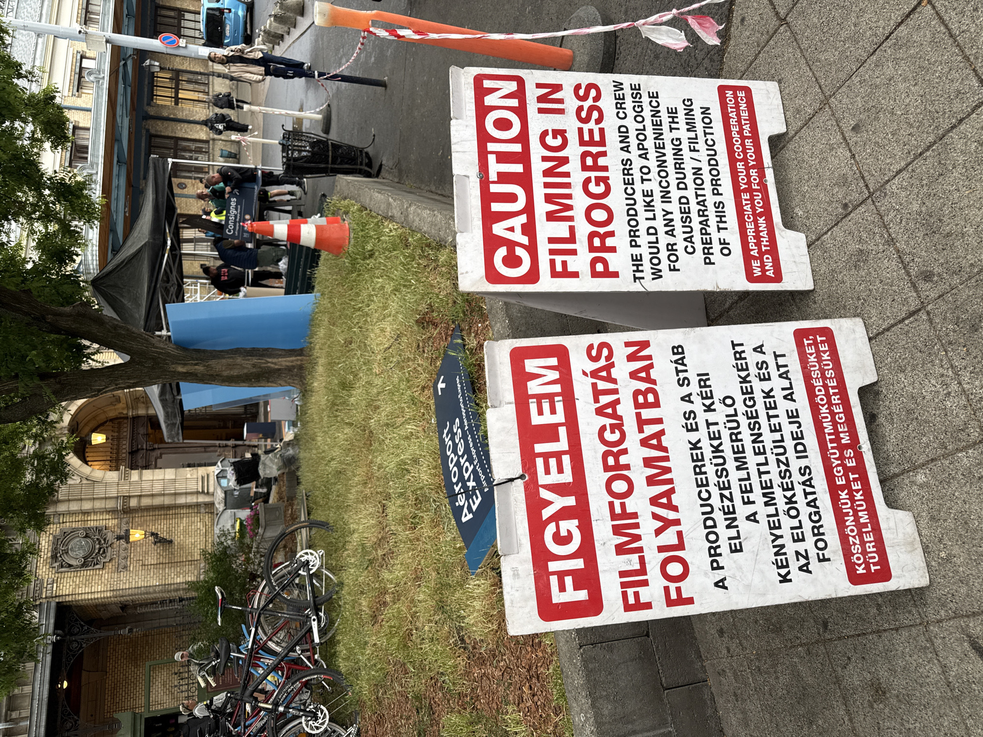 The image depicts a film set in Budapest, Hungary. Two signs, one in English and one in Hungarian, warn of filming in progress. There is a sign for an airport express. A group of people stands under a dark tent structure, near an orange traffic cone. A rack of bicycles is parked nearby. The backdrop includes a building with visible architectural details, including arched entrances and ornate decorations.