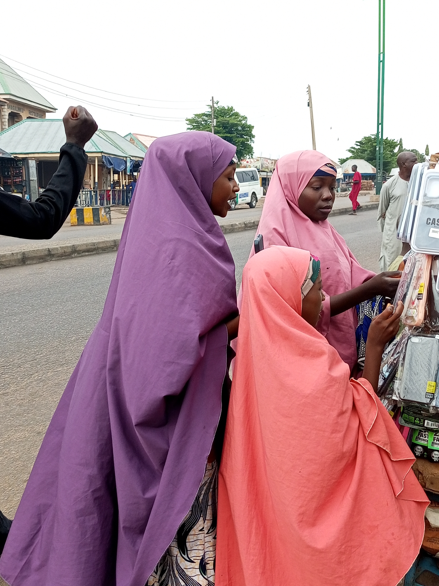 In Tempure, Nigeria, three young women dressed in traditional Islamic attire, or hijabs, are browsing a street vendor's stall. The woman on the left wears a vibrant purple hijab, while the woman in the middle and the woman on the right wear pink hijabs. The woman on the right is holding a phone case and examining it closely, suggesting she is considering a purchase. The stall itself is filled with various phone cases, displayed on a rack. In the background, a man in a light-colored garment walks away from the stall, and a street scene unfolds with buildings, a road, and a few parked vehicles, including a white van. The overall atmosphere appears to be a typical marketplace in a Nigerian city.