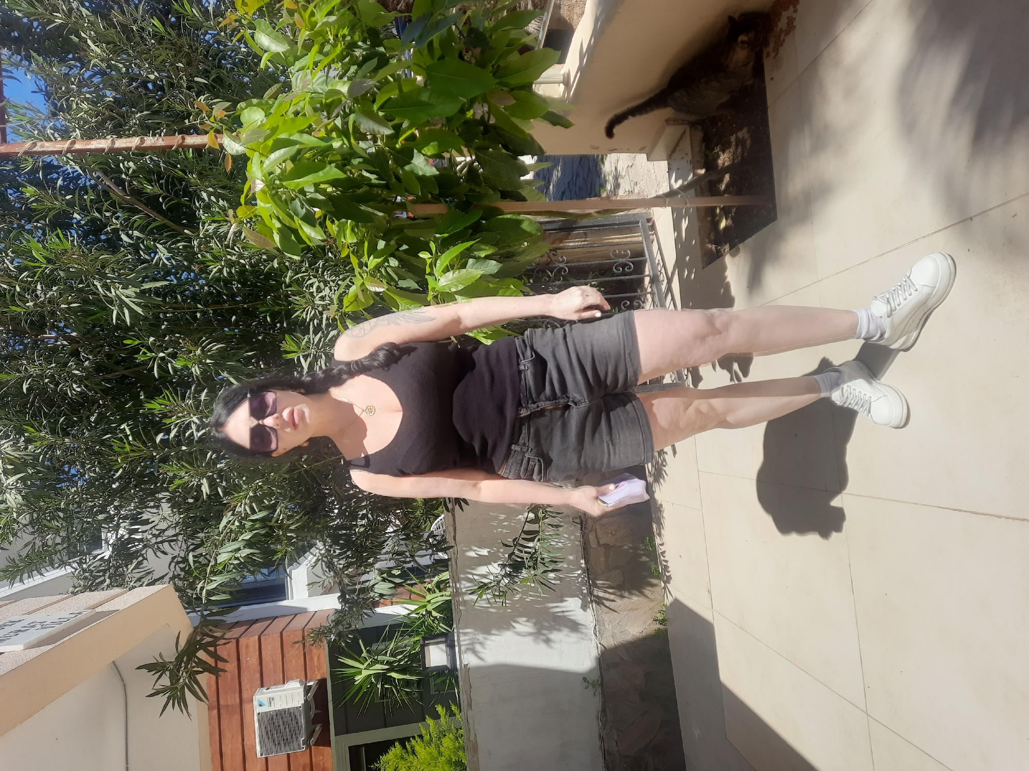 An outdoor, sunlit scene in Kemer, Turkey, features a woman standing on a light-colored tiled patio, surrounded by lush green foliage. The image captures a bright daytime setting, indicated by strong sunlight and pronounced shadows cast by subjects and plants.
The woman, with dark hair, stands facing the camera. She wears a black tank top, dark shorts, white sneakers, and white socks. Sunglasses obscure her eyes, and a necklace is visible around her neck. Tattoos adorn her left arm, and she holds a small, light-colored object in her left hand. Her posture is relaxed, and her expression is neutral.
In the upper right portion of the frame, a dark-furred cat is partially visible, nestled among bushes and shadows on an elevated surface or ledge. It appears to be looking towards the right.
The background is dominated by dense green trees and bushes, characteristic of a Mediterranean landscape. To the woman's left, parts of buildings are visible; one structure features a brown wooden facade with a white air conditioning unit installed beneath a window. Another nearby building displays a partially visible sign that reads "NO SMOK...", likely indicating "NO SMOKING". A metal railing is also present in the mid-ground. Clear blue sky can be seen through gaps in the dense foliage, highlighting the sunny weather.