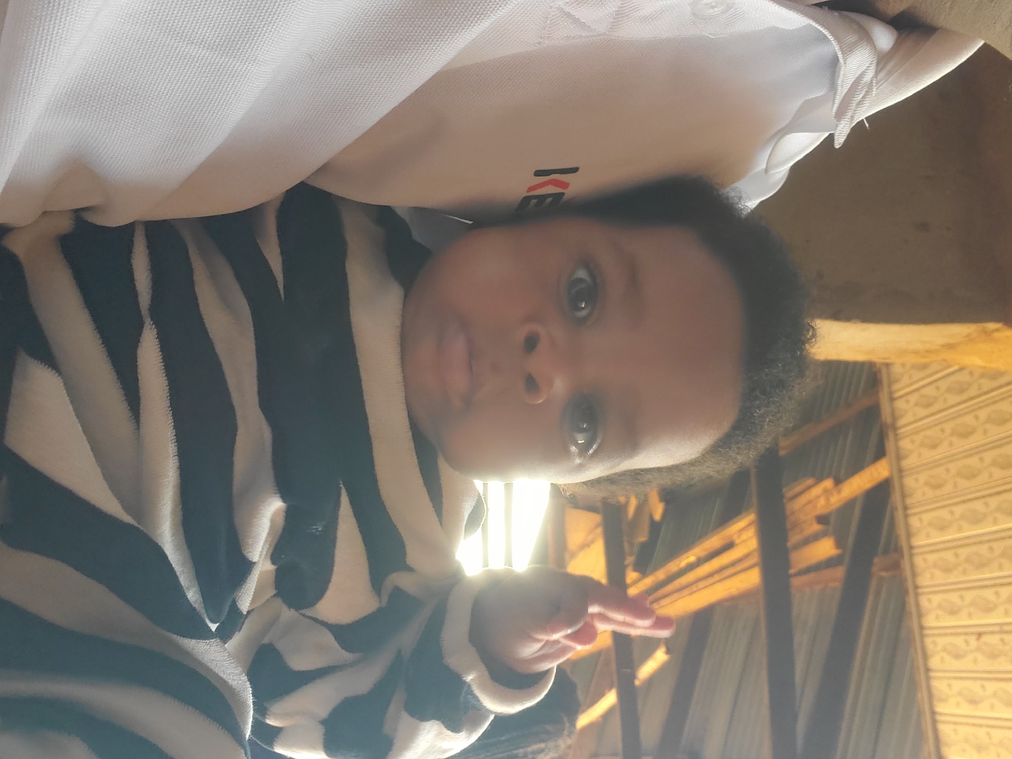 A baby with dark curly hair is lying on its back. It is wearing a zebra-striped outfit. The baby is looking upwards and slightly to the right, with its mouth slightly open and tongue protruding slightly. One hand is raised in the air, fingers extended. Above the baby is a light-colored garment with the letters "KE" visible. In the background is a ceiling with support beams and patterned paneling. The location is Ukwu-Abwa, Nigeria.