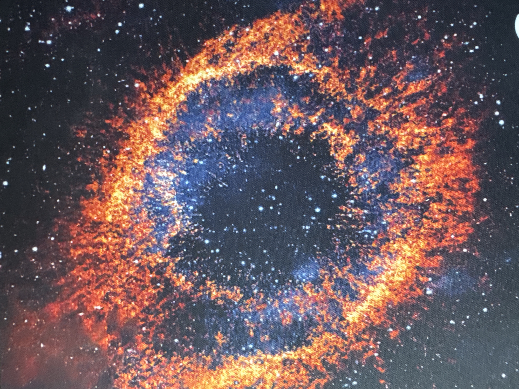 The image is a close-up, detailed view of a nebula, likely an artistic representation of a celestial object. It displays vibrant colors, predominantly fiery oranges and deep blues, interspersed with speckles of white that represent stars. The texture of the image suggests it might be a photograph of a printed material, such as a poster or fabric, due to visible grain or weave patterns. The nebula itself is ring-shaped, with a dark central void. The composition is abstract and cosmic. The location is unknown.