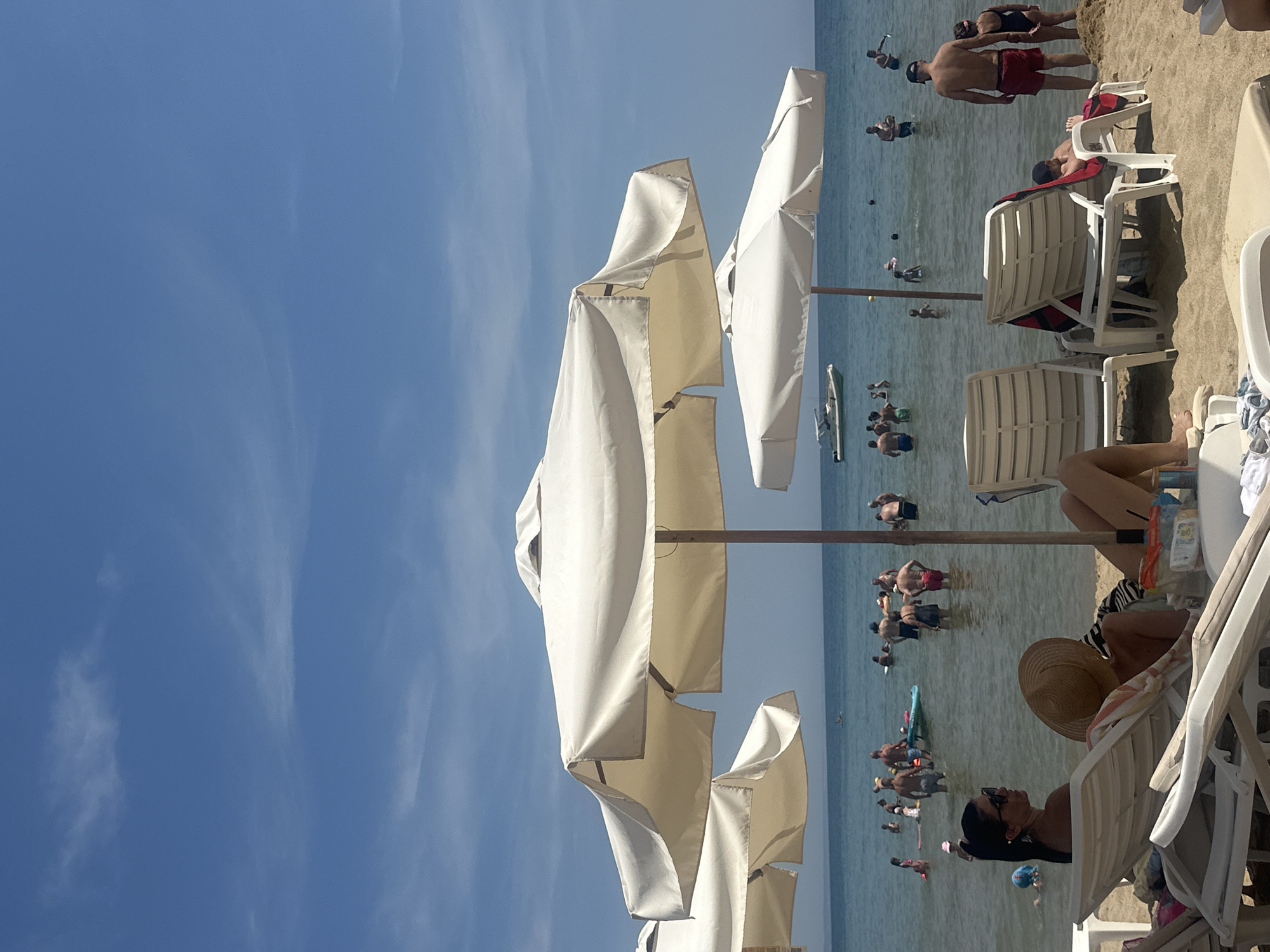 The image depicts a crowded beach scene under a bright blue sky with scattered white clouds, likely in the city of Varna, Bulgaria, given the context. In the foreground, several white beach chairs and umbrellas are set up on a sandy shore. Two women are seated on chairs, with one wearing sunglasses and the other a straw hat.
The middle ground shows the shallow, clear water filled with numerous people swimming, wading, and playing. Some individuals are standing, while others are partially submerged. A blue inflatable raft is visible among the swimmers. In the background, a small white boat is sailing on the sea, with the horizon line separating the water from the sky. The overall atmosphere is one of a lively summer day at a popular beach resort.