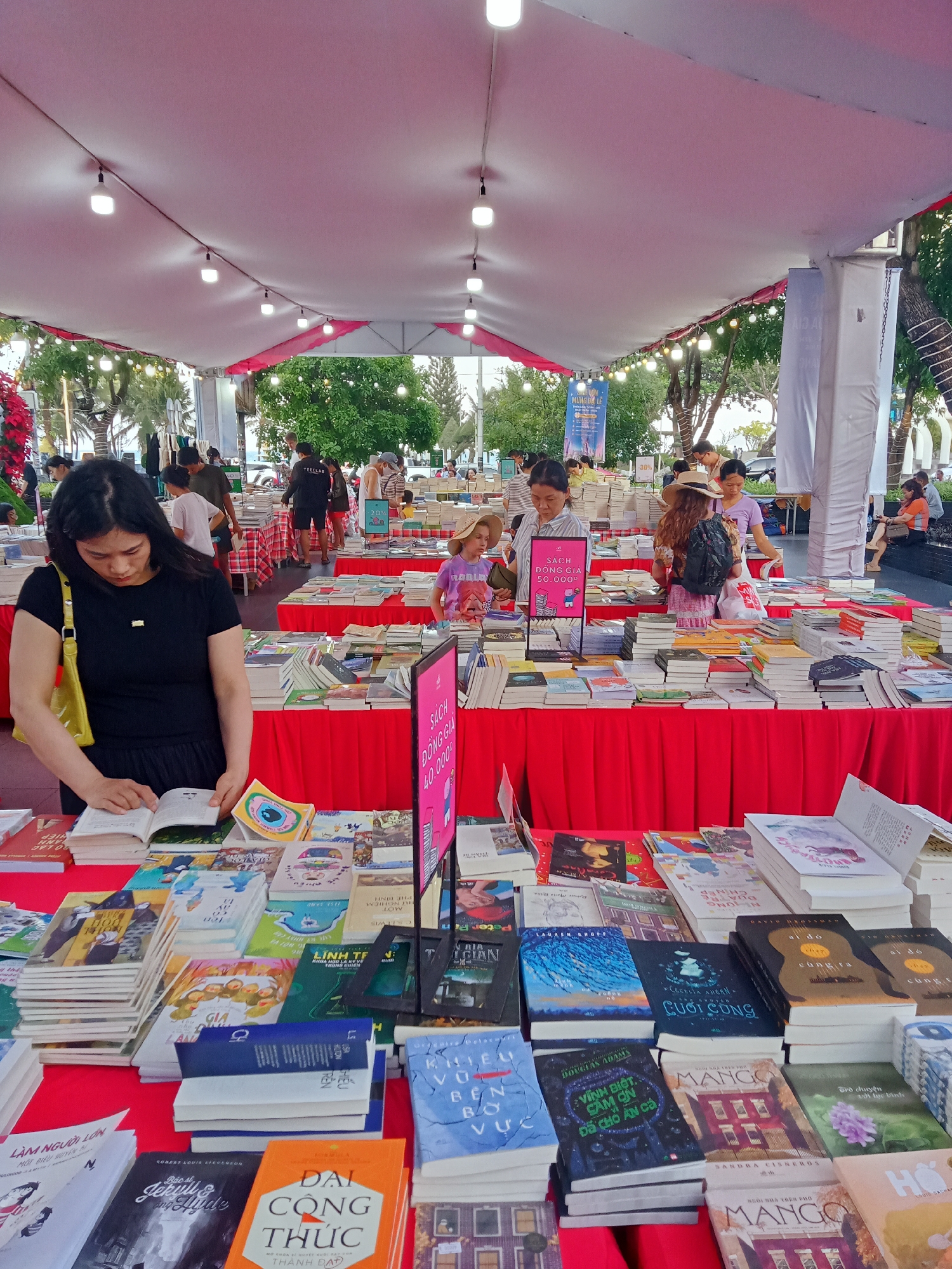The image shows a bustling outdoor book fair, likely in Nha Trang Ward, Vietnam, based on the language of the book titles and signage. A large tent structure provides shade for multiple book stalls, each covered with red tablecloths. People of various ages are browsing the books on display. In the foreground, a woman in a black dress and yellow bag is intently examining a book. Other individuals, including a child in a straw hat and several adults, are perusing the selections at different stalls. Signs advertising book prices ("SACH DONG GIA 40.000" and "SACH DONG GIA 50.0000") are prominently displayed. The scene is brightly lit, and trees are visible in the background, suggesting a location near a park or outdoor area. The books are stacked in piles on the tables, covering a wide variety of genres and topics.