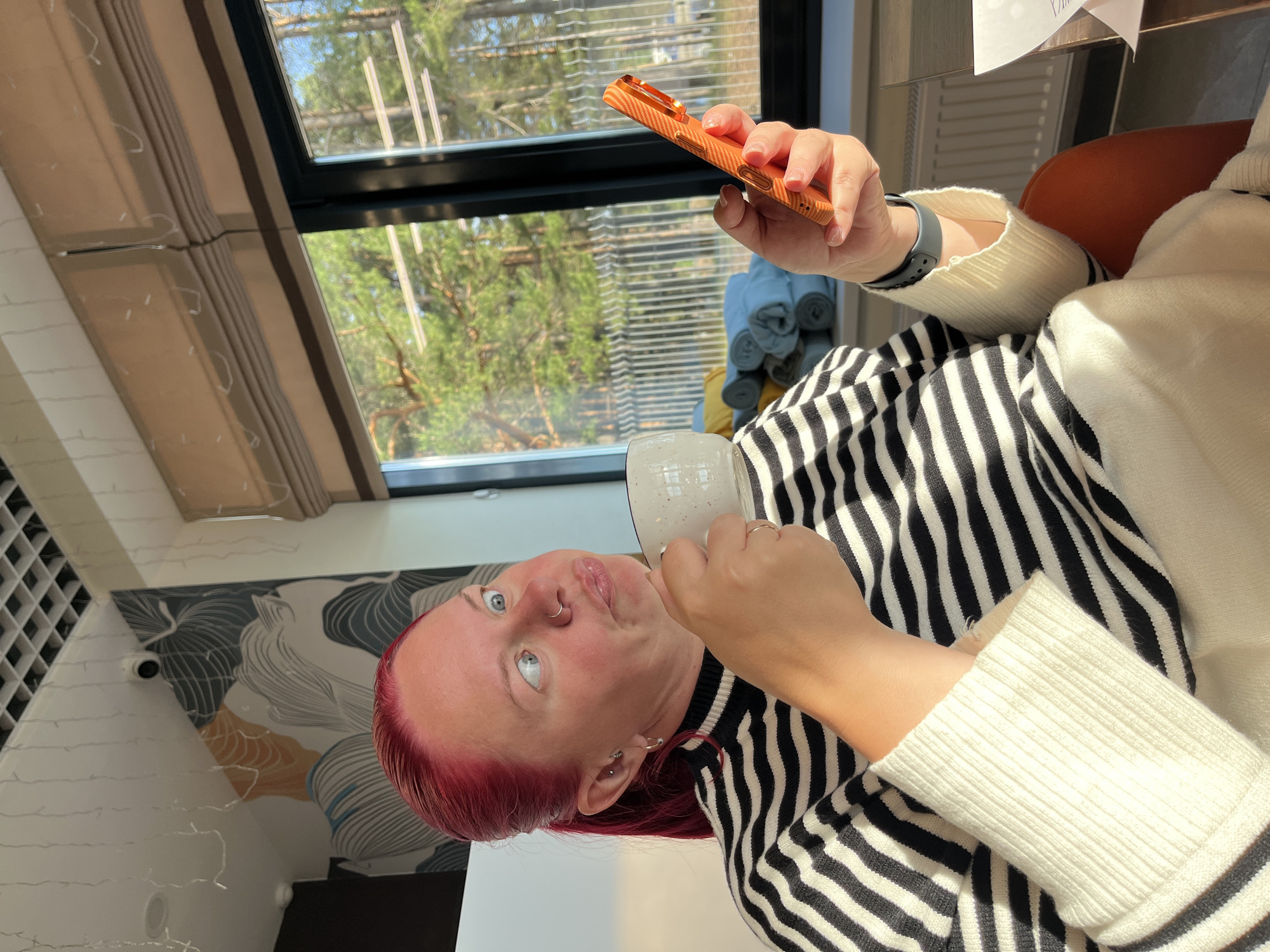 A person with vibrant red hair, light skin, blue eyes, a nose ring, and ear piercings is depicted in a medium close-up shot. They are wearing a black and white striped long-sleeved top. With their right hand, they hold a white, speckled ceramic mug to their lips, while their left hand holds an orange or copper-colored smartphone with a textured case. A dark-banded smartwatch is visible on their left wrist. The person's gaze is directed upwards and to the right, with a slightly open mouth, suggesting focus or surprise.
The setting is indoors during the daytime, brightly lit by natural light. A large window, likely a skylight, is visible in the background, looking out onto lush green trees and a clear blue sky, indicating sunny weather. Brown blinds are partially drawn on the window. To the right of the person, stacked rolled textiles in blue and yellow are visible. The left side of the background features a white wall with a modern, abstract pattern of swirling lines in gray, blue, and orange. A grid-like structure, possibly a wine rack or shelving, is on the far left wall, along with faint fairy lights. A small white security camera is mounted on the wall near the patterned wallpaper. Part of an orange or brown chair back and a white table surface are also visible. The scene captures a moment of leisure, likely in a home or cafe, in Chaikovsky, Russia. No visible text is discernible.