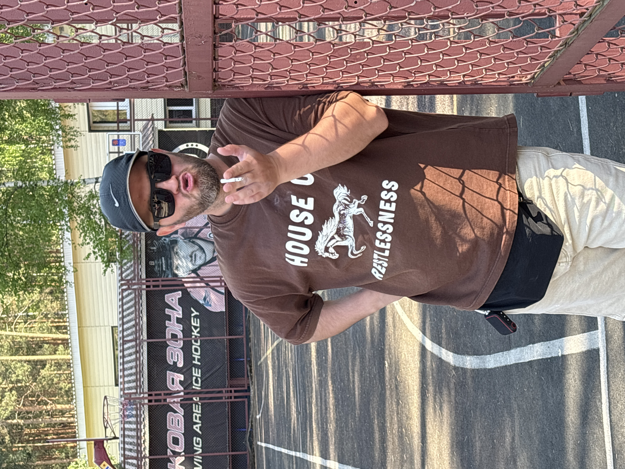 A man wearing a brown t-shirt, tan pants, a black Nike baseball cap, and sunglasses is standing outside. He is holding a lit cigarette in his right hand and has a black bag around his waist. He stands next to a chain-link fence. Behind the fence is a basketball court with white lines. Further back, a large banner is visible that reads "КОВАЯ ЗОНА ICE HOCKEY" (KOVAYA ZONA ICE HOCKEY), which identifies the location as Chaykovsky, Russia. There is a building in the background with trees visible on the left side of the frame.
