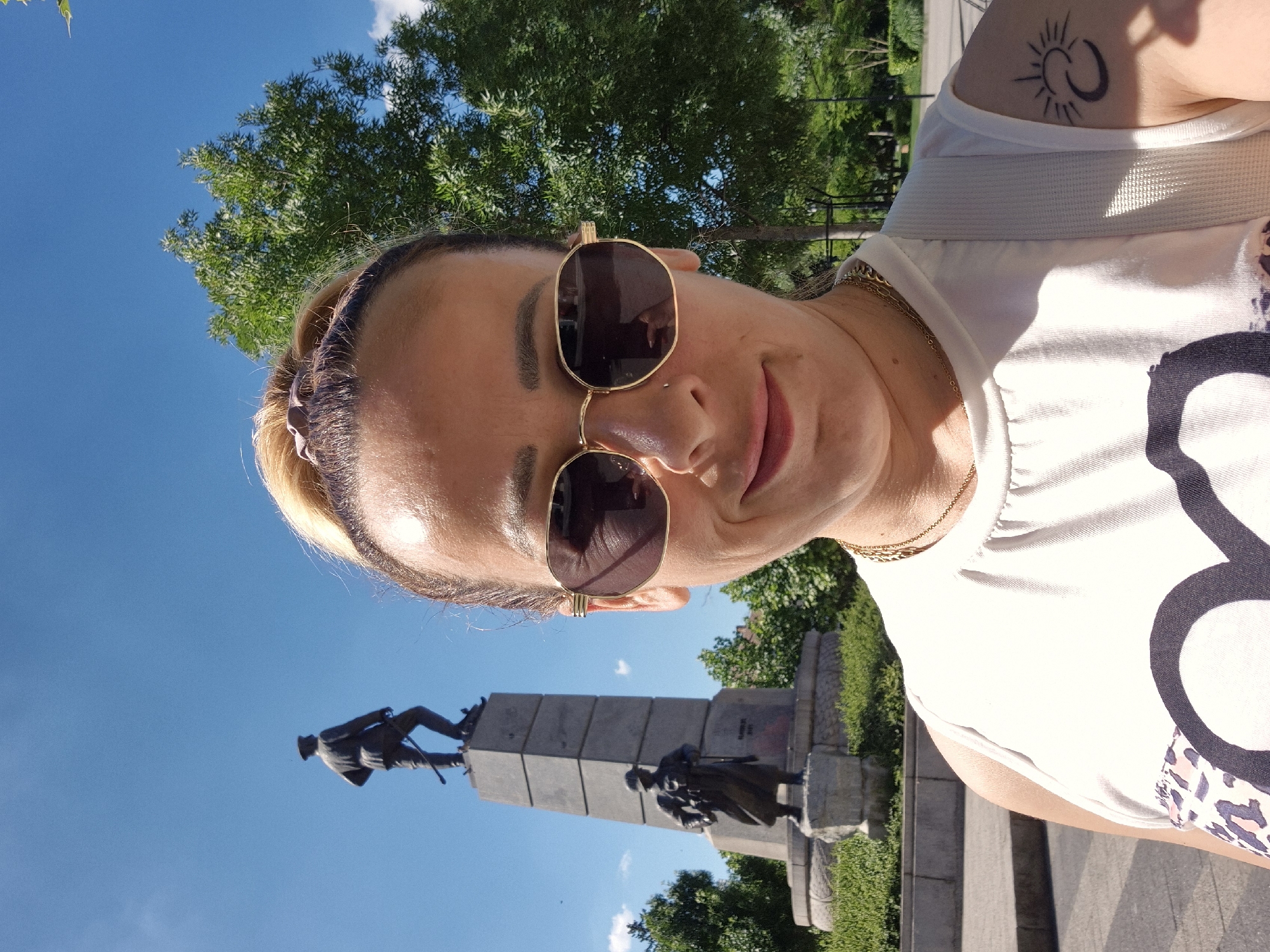 A woman is taking a selfie outdoors on a sunny day in front of a monument. She has blonde hair pulled back and is wearing sunglasses and a white tank top. A tattoo of a sun and moon symbol is visible on her left arm. Behind her, a tall, stone monument with two statues stands against a bright blue sky. Lush green trees and park landscaping surround the monument. The ground in the foreground appears to be paved.