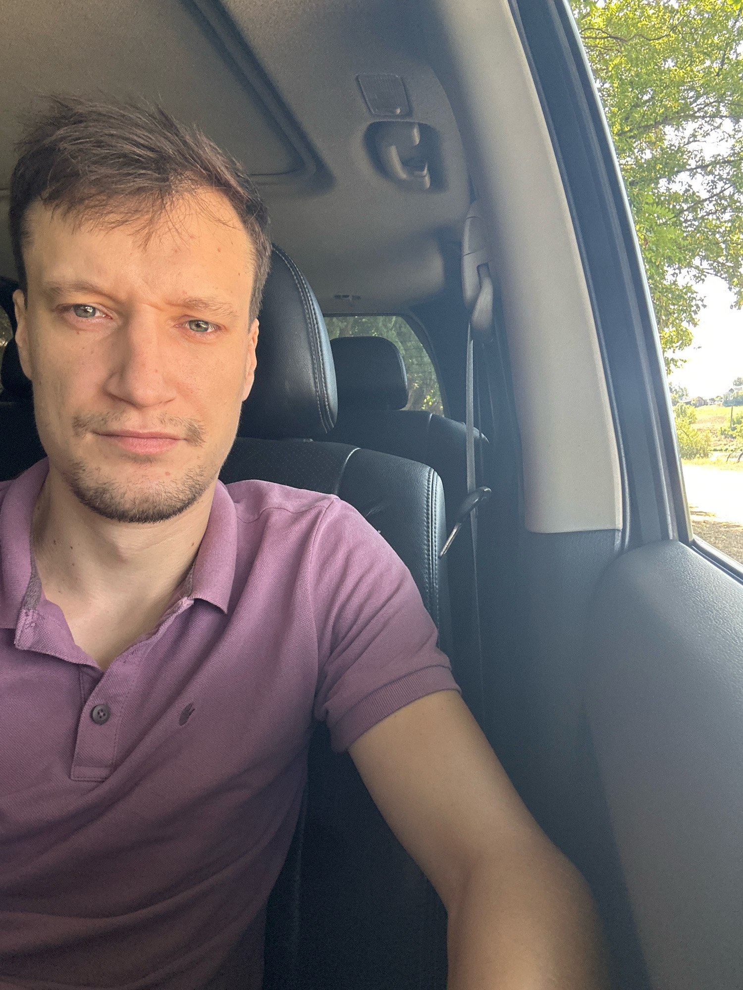 A young man with light skin, brown hair, and light-colored eyes is taking a selfie from the driver's seat of a car. He is wearing a purple polo shirt. His left arm is resting on the center console. The interior of the car is visible, including black leather seats, a sunroof, and a grey interior trim. Outside the car, trees and a rural landscape with houses are visible. The time of day appears to be daytime, with sunlight filtering into the car. The specific location in Makiv, Ukraine is not discernible from the image.
