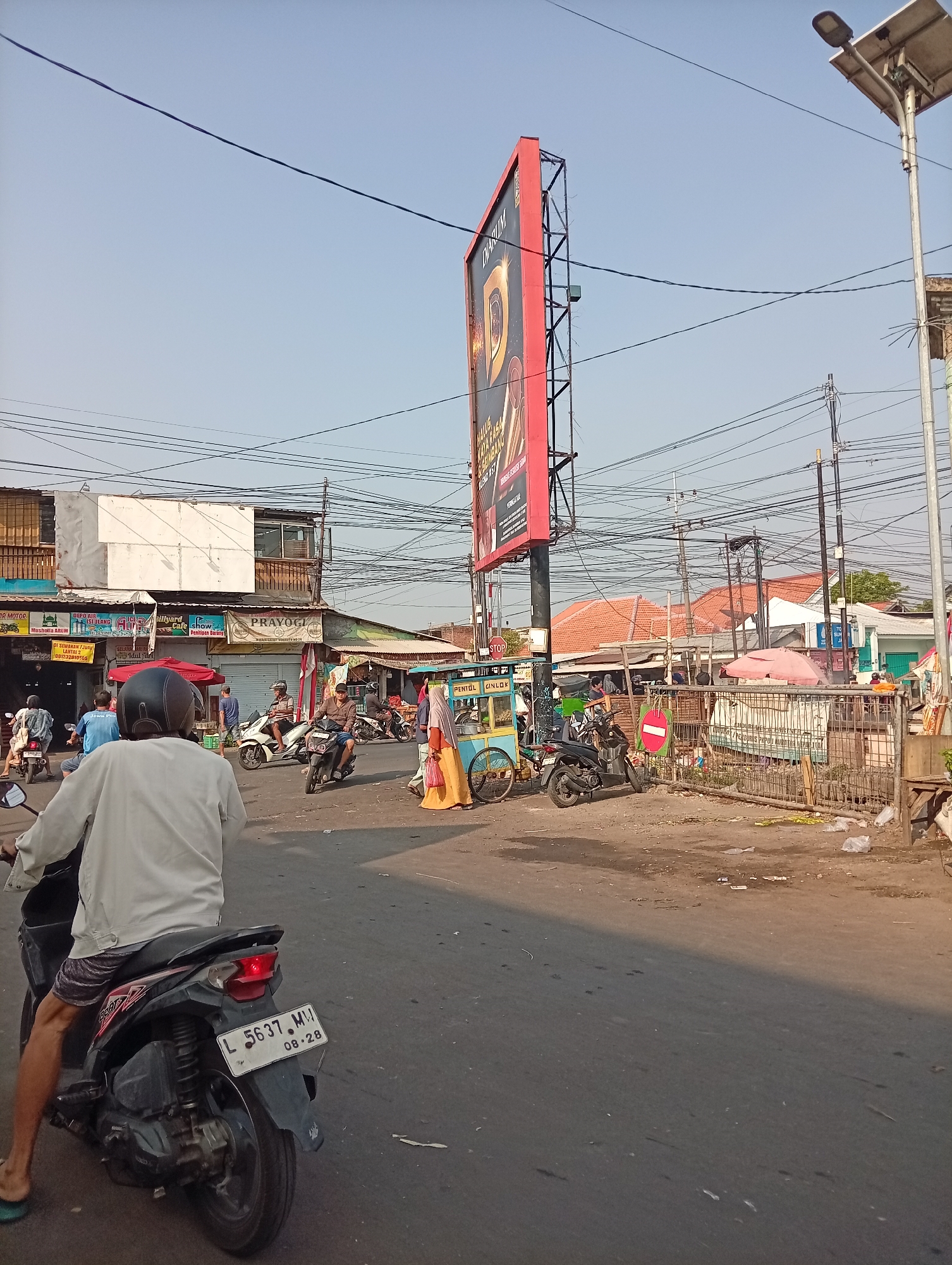 The image depicts a street scene in Larangan, Indonesia. In the foreground, a person is riding a dark-colored motorcycle with a license plate reading "L 5637 MW 08-28". They are wearing a light-colored jacket and patterned shorts. The motorcycle has a red brake light illuminated. The street is paved and appears to be busy with other motorcycles and pedestrians.
To the left, a row of shops and buildings lines the street. One building has a white facade and a second story with balconies. Signs are visible above the shops, including one that reads "PRAYOGI".
In the background, a tall billboard with a red frame displays an advertisement. A street light with a solar panel is mounted on a pole to the right of the billboard. More buildings with orange tiled roofs are visible further back, along with numerous electrical wires crisscrossing overhead. A cart with a yellow base and a blue top, possibly a food cart, is parked near the shops. People are seen walking and riding motorcycles on the street and sidewalk. The overall environment suggests a bustling urban or market area.