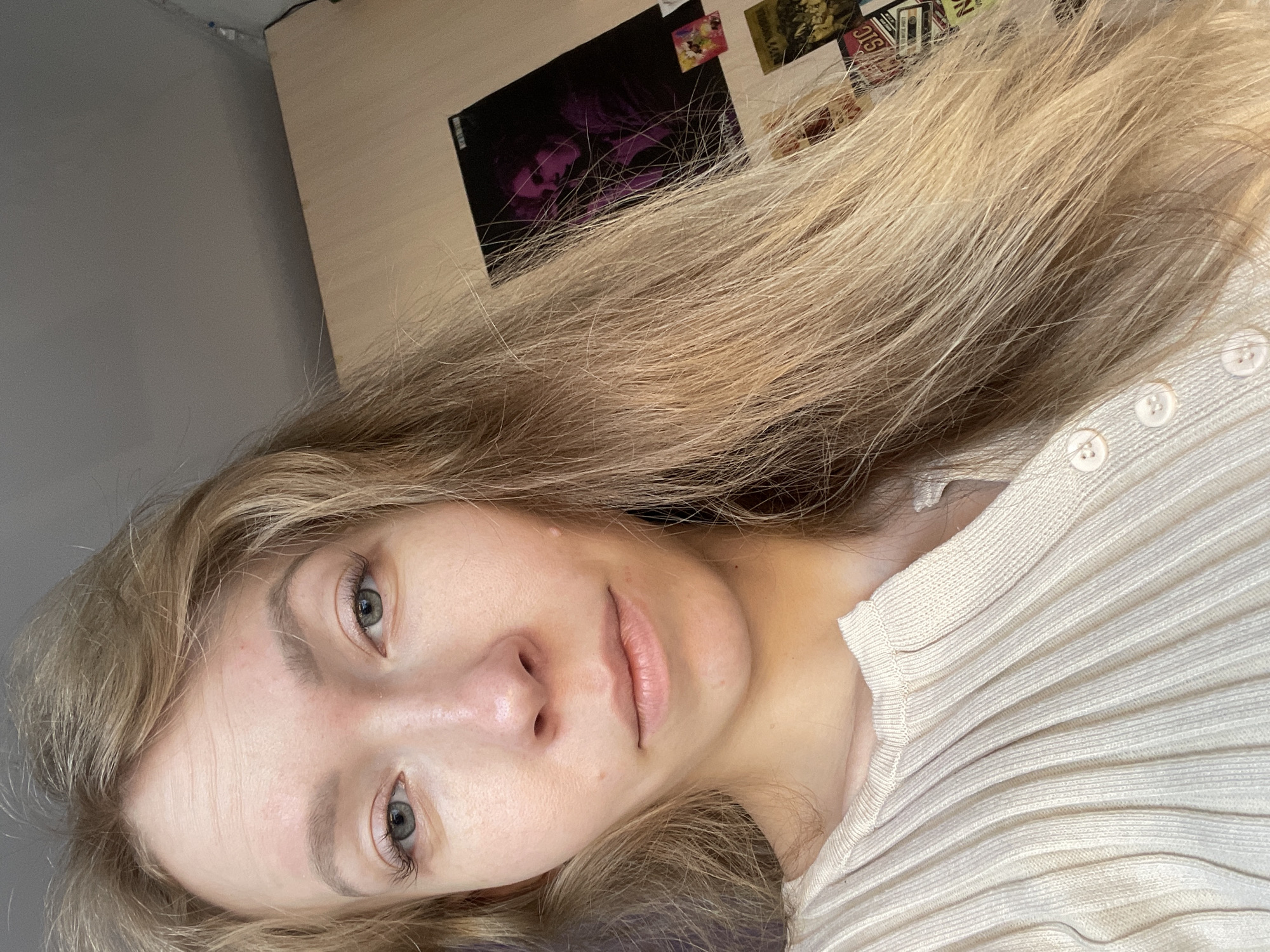 A selfie photograph shows a young woman with light blonde hair and fair skin. She is wearing a ribbed, light-colored top with buttons at the neckline. Her eyes are light-colored, and she is looking directly at the camera. The background includes a light-colored wall with several pictures or postcards attached. A gray wall is also visible. There are no indicators for determining the location of Siva, Russia in the image.