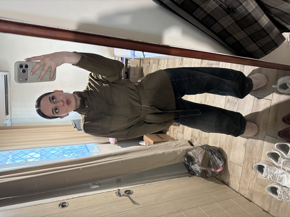 The image depicts a mirror selfie of a young female inside what appears to be a residential interior in Sofia, Bulgaria. The subject, a female with dark hair pulled back, is wearing an olive-green belted jacket with a high collar and front pockets, dark, loose-fitting jeans, and light pink slip-on slippers. She holds a light-colored smartphone with an ornate case in her right hand to capture the reflection, looking towards her left with a pouting expression.
The environment is a room with light wooden flooring. To the left of the subject, a light-colored wooden door with a silver handle is partially visible, next to a beige curtain covering a window with visible blue-framed security bars or a grille. A black plastic trash bag rests on the floor near the door. Multiple pairs of shoes, including white sneakers, are arranged on the floor in the foreground. In the background, an air conditioning unit is mounted on a light-colored wall above the window area, and a cluttered surface, possibly a desk or dresser, with papers and a brown cardboard box is discernible. A thick, reddish-brown wooden frame defines the right edge of the mirror, behind which a plaid shirt is partially visible.