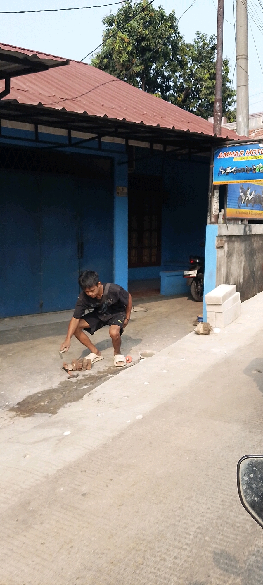 A young male is crouched on a concrete surface in front of a blue building. He is wearing a black t-shirt and black shorts, with white flip-flops on his feet. He is holding a trowel in his right hand and appears to be working with a stack of bricks in front of him. There are also some scattered objects on the ground around him, including a small pink toy and a few stones. Behind the boy is a blue building with a red corrugated metal roof and a dark-colored door. A motorcycle is parked to the right of the building entrance. A sign for "AMMAR MOTOR" is visible on the wall of the building, indicating a motorcycle shop. The scene appears to be on a street in Depok, Indonesia, with a sidewalk and a road visible.