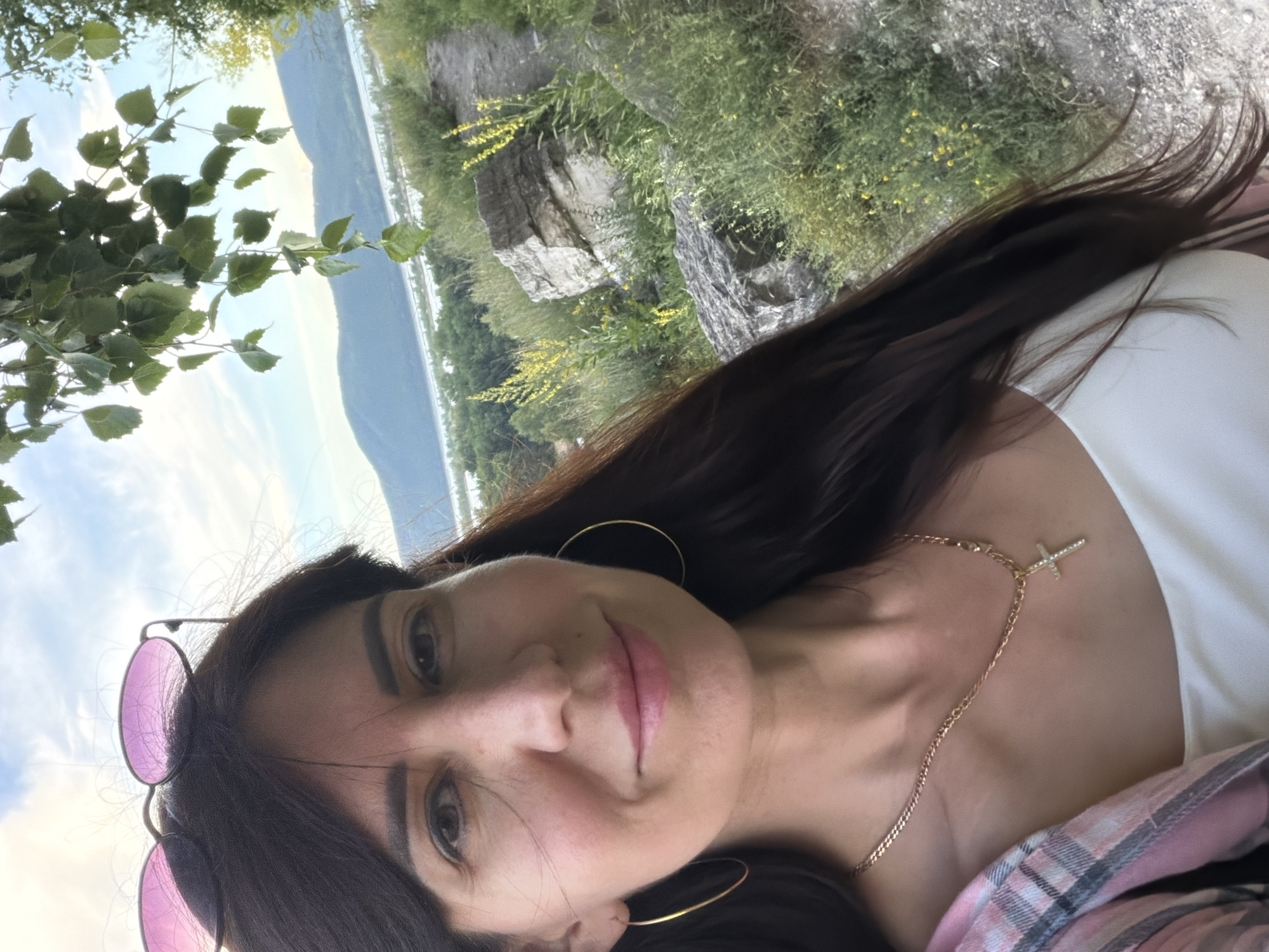 A woman is taking a selfie outdoors. She has long, dark hair and is wearing pink-tinted sunglasses perched on her head, large hoop earrings, a gold chain necklace with a cross pendant, and a white top. She is also wearing a pink plaid shirt partially visible. The background shows a natural landscape with trees, rocks, and a body of water in the distance, possibly the Volga River, suggesting a location in or near Volzhskiy, Russia. The sky is cloudy.