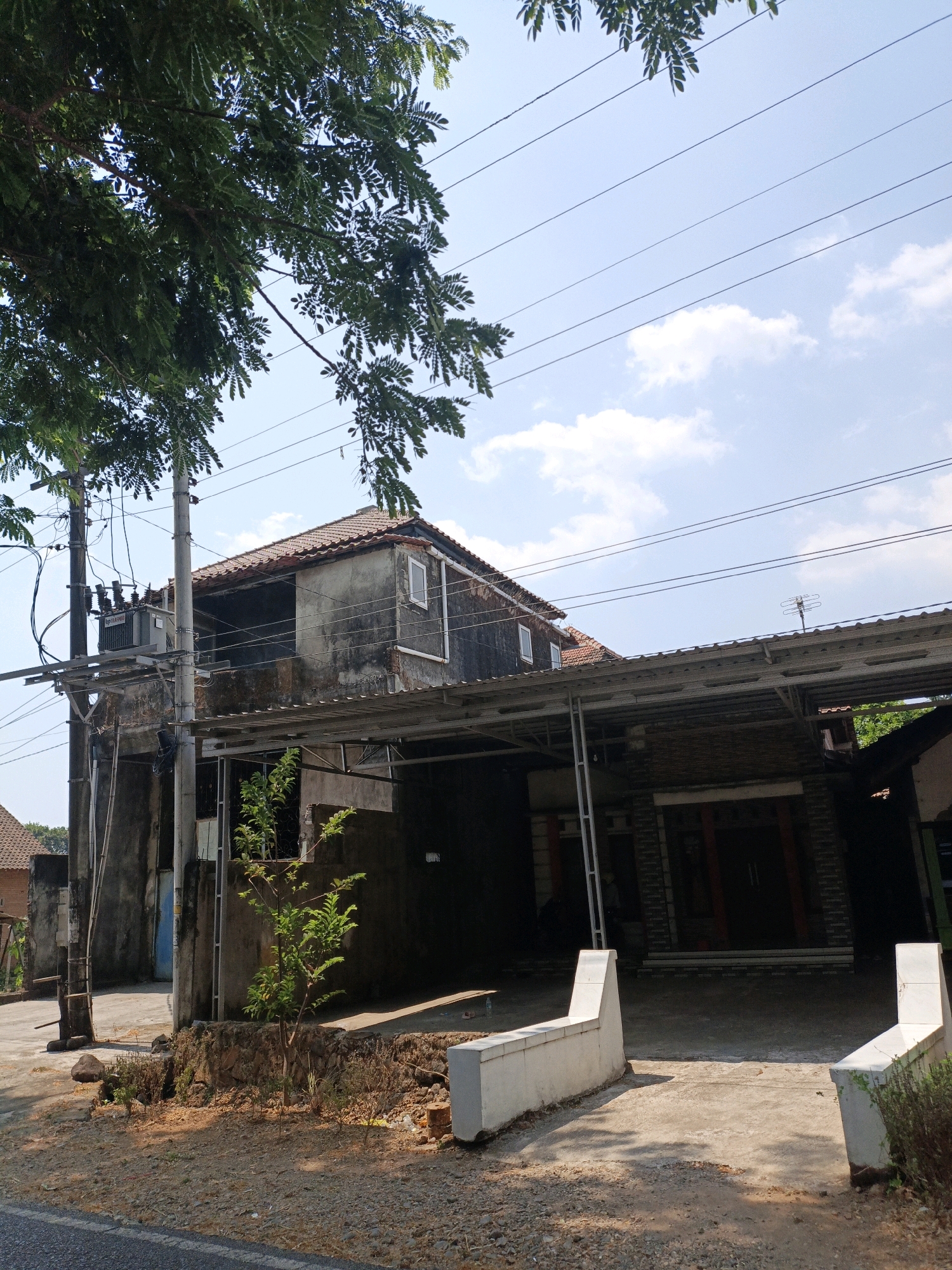 The image shows a two-story house with a weathered exterior, a tiled roof, and several windows. A carport extends from the front of the house. Power lines and a transformer are visible on the left side of the house. A small tree and various plants are growing in front of the house, which is situated on a dirt and gravel area next to a paved road. The sky is blue with some clouds. The scene appears to be in a residential area in Cikal, Indonesia. There are no people or animals visible in the image.