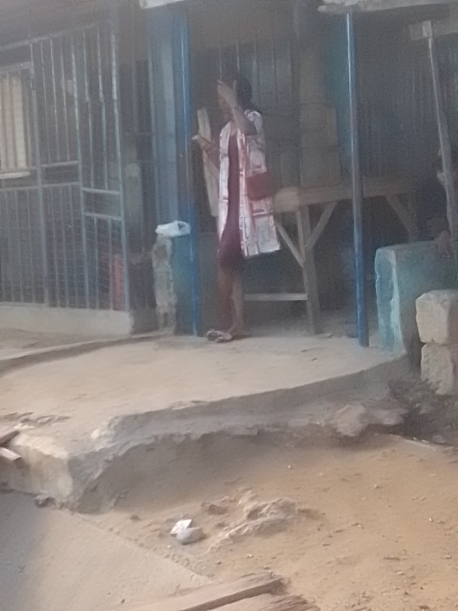 The image captures a scene in what appears to be a bustling street market or commercial area in Galadima, Nigeria. In the foreground, a sandy, uneven ground is visible, with some debris and a small concrete structure. In the mid-ground, a woman is standing in front of a covered stall. She is wearing a patterned dress and holding what looks like a red bag. Behind her, there is a wooden table or counter. The background features a metal grate structure, possibly part of a building or stall. The overall impression is one of a typical day in a Nigerian urban setting, with a focus on a local vendor and her merchandise.