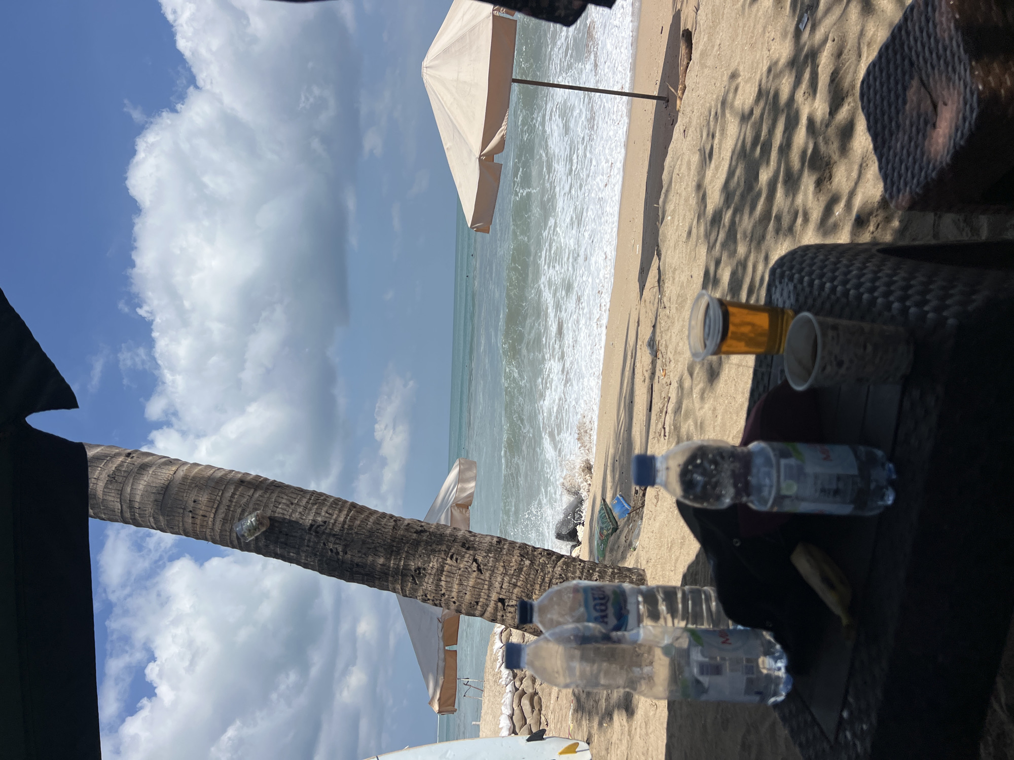 In Kuta, Indonesia, a sunny day unfolds on a beach. A tall, rough-barked palm tree stands prominently in the foreground, partially obscured by a black awning. In front of the tree, a woven table holds several objects: three plastic bottles of water, a black cap, a banana, and two cups, one containing a dark beverage and the other filled with a golden-colored liquid, likely beer. Beyond the table, the sandy beach stretches towards the ocean, where gentle waves crash onto the shore, creating white foam. Two light-colored beach umbrellas are set up, one closer to the water and another further back. In the distance, a few figures can be seen in the sea, suggesting people swimming or surfing. The sky is a bright blue, dotted with fluffy white clouds.
