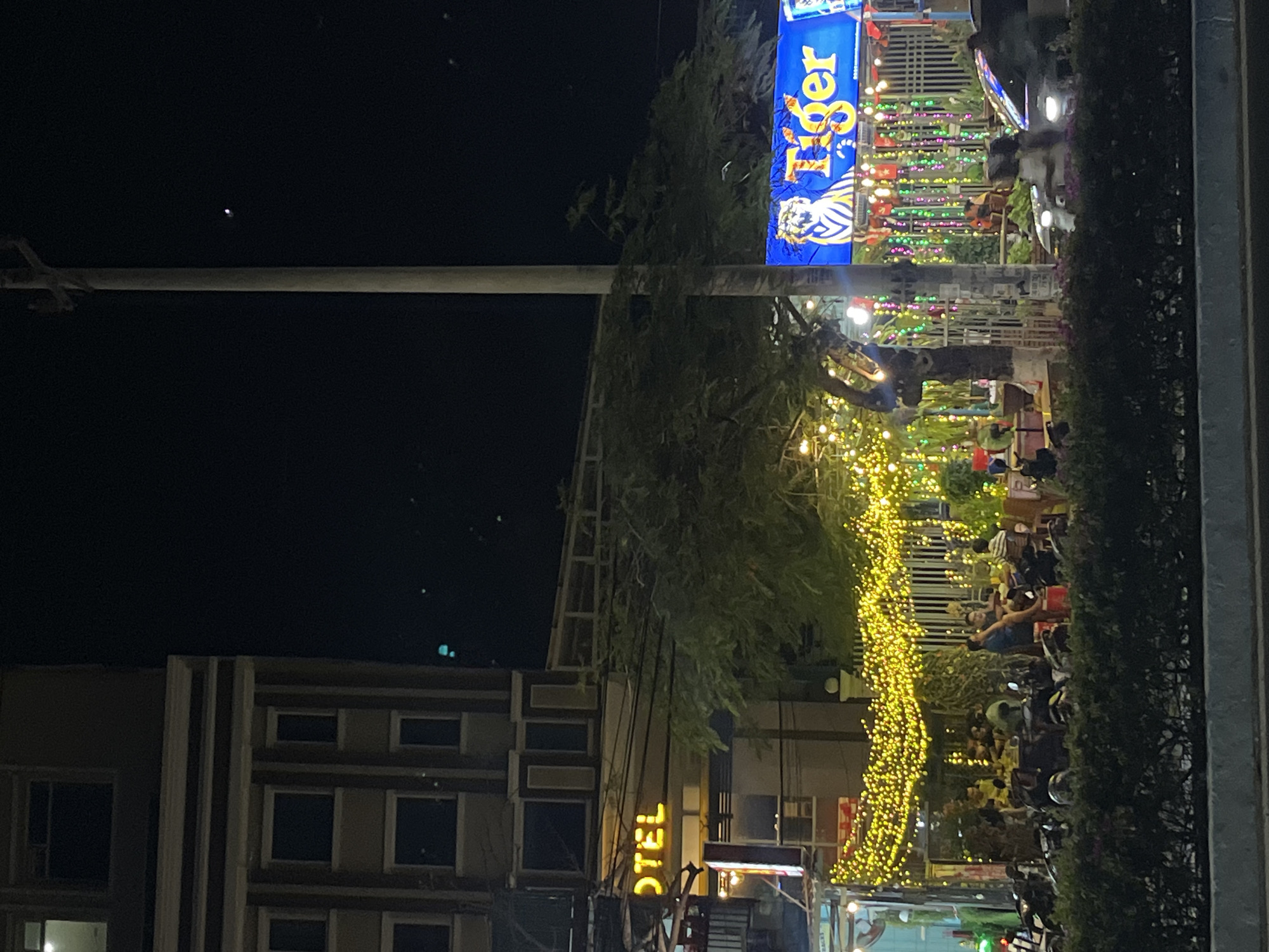 At night, the Bac Nha Trang Ward in Nha Trang, Vietnam is lively with people sitting at outdoor establishments. A Tiger beer sign is displayed. There are strands of yellow lights. Parked motorbikes fill the sidewalks. A building with a sign that says “Hotel” is partially visible. A few utility poles are visible against the dark sky.
