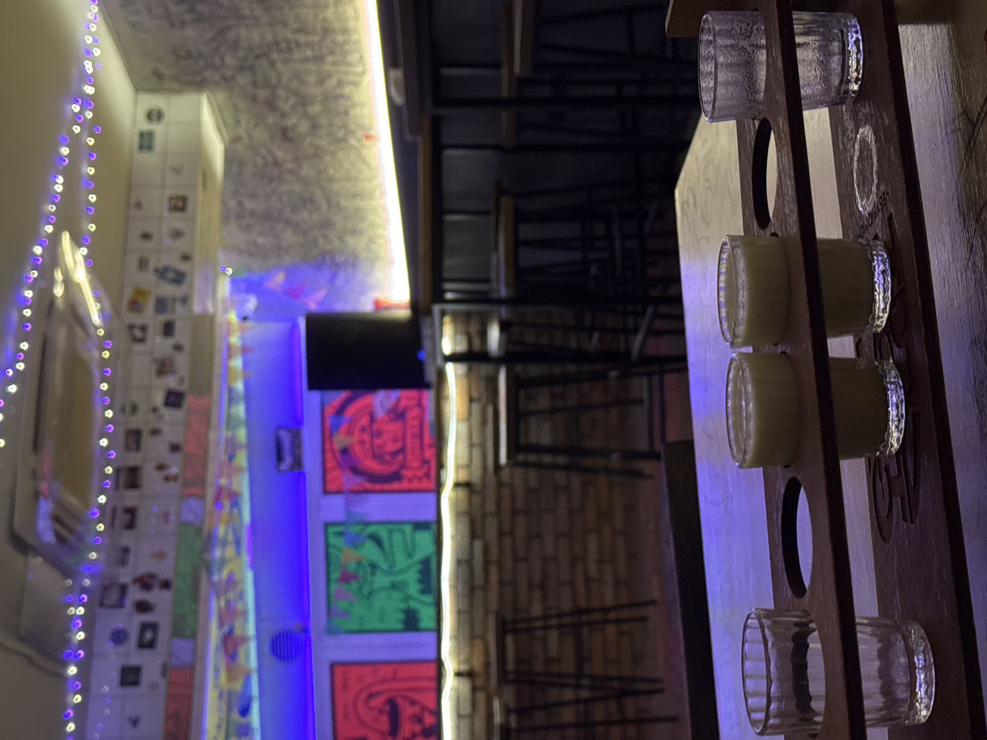 The image depicts a dimly lit interior, possibly a bar or pub, with a focus on a wooden tray holding four small glasses on a table. Two of the glasses are filled with a pale yellow liquid, while the other two are empty and appear to be made of clear, textured glass. In the background, a brick wall serves as a backdrop for several framed pictures and colorful artwork displayed on the wall above. Stools are arranged in front of a counter with neon blue lighting. String lights with purple and white bulbs are visible along the ceiling. The overall ambiance suggests a casual, possibly artistic or bohemian setting.